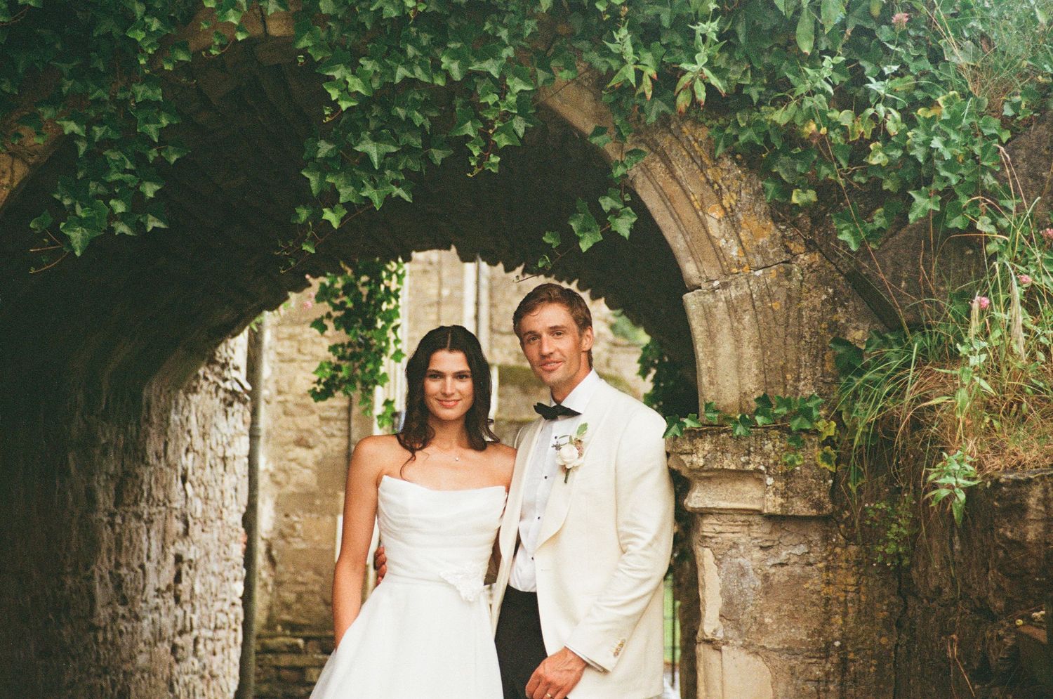 Bride and groom posing together under lush ivy-draped historic stone archway.