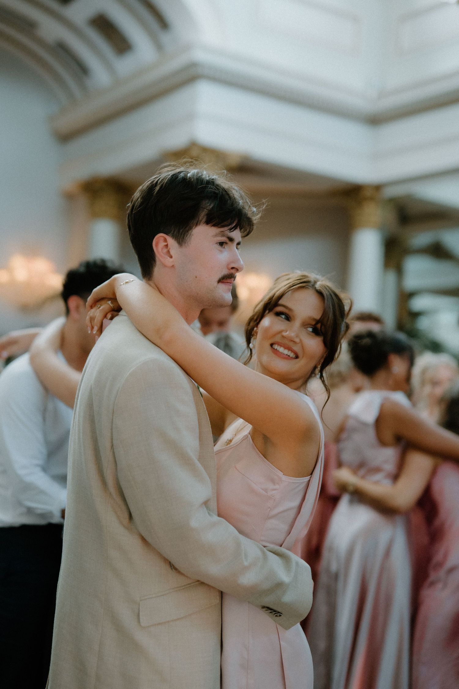 Bride smiling while dancing with a guest in a beige suit during the wedding reception in an elegant grand ballroom.