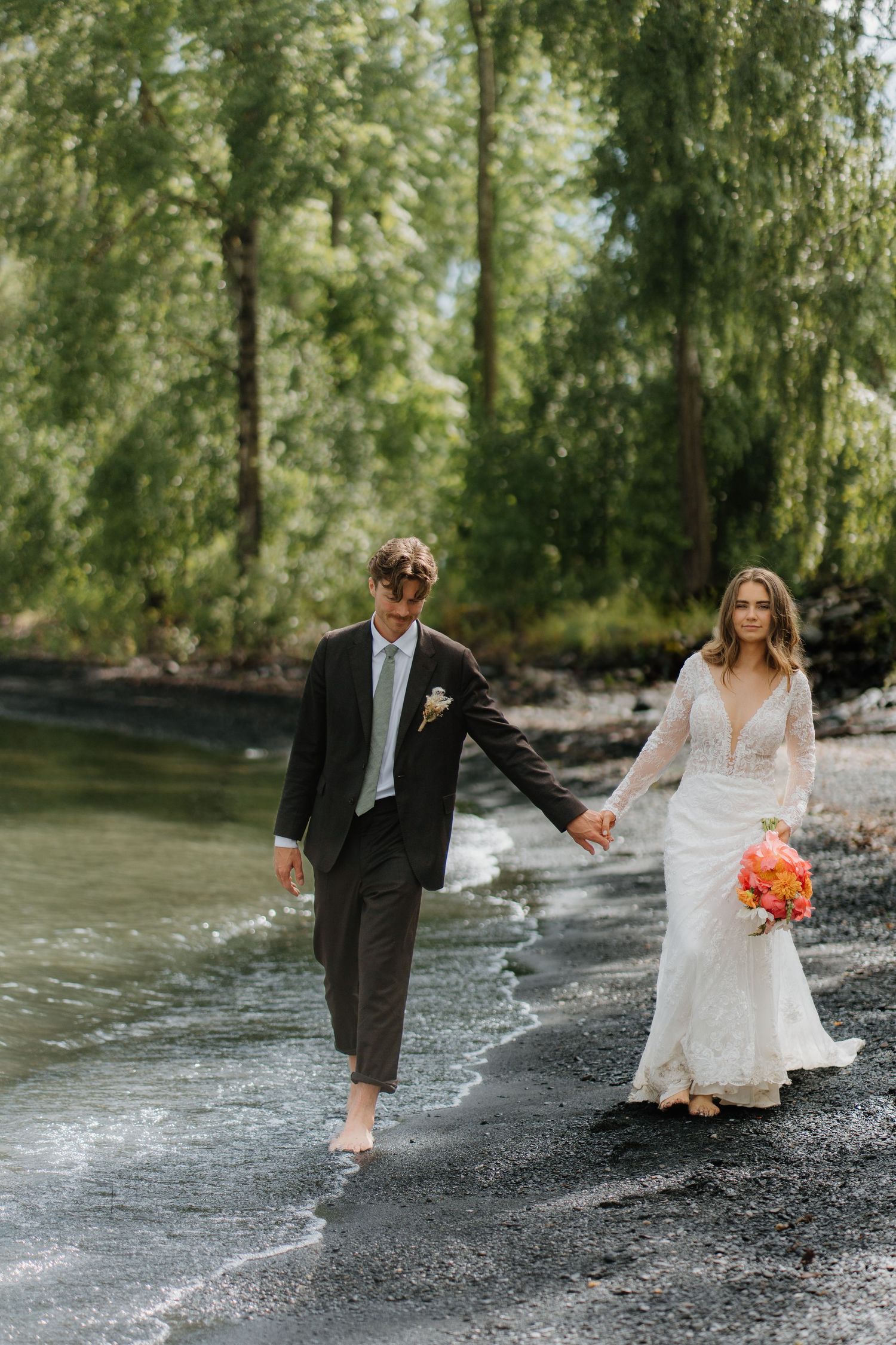 Bride and groom walk barefoot along shoreline holding hands, bride carries bright floral bouquet.