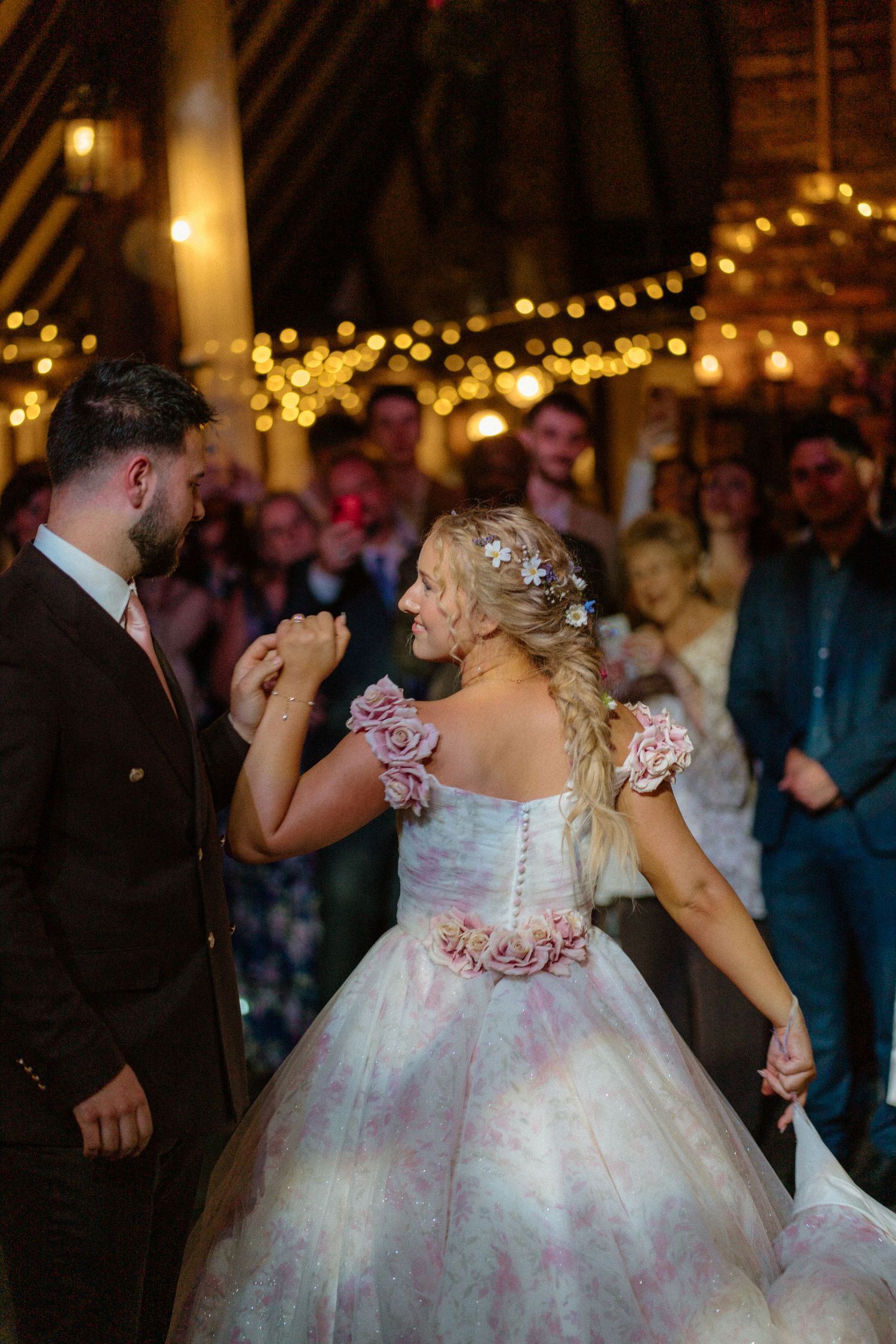 Bride in pink floral gown holds grooms hand during first dance surrounded by guests in a fairy-lit barn.