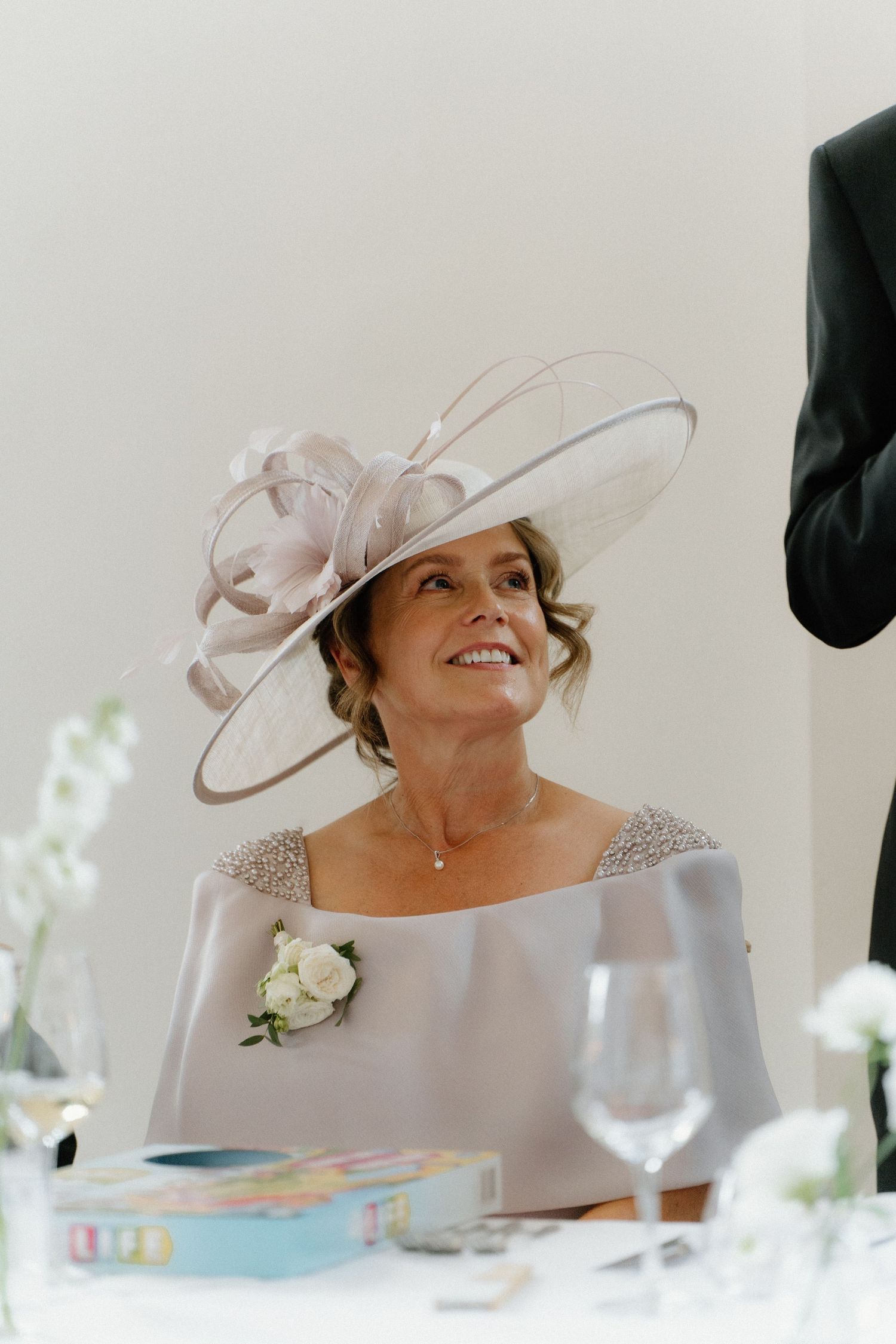 Elegant mother of the bride wearing a blush fascinator hat and grey gown with floral corsage at a wedding.