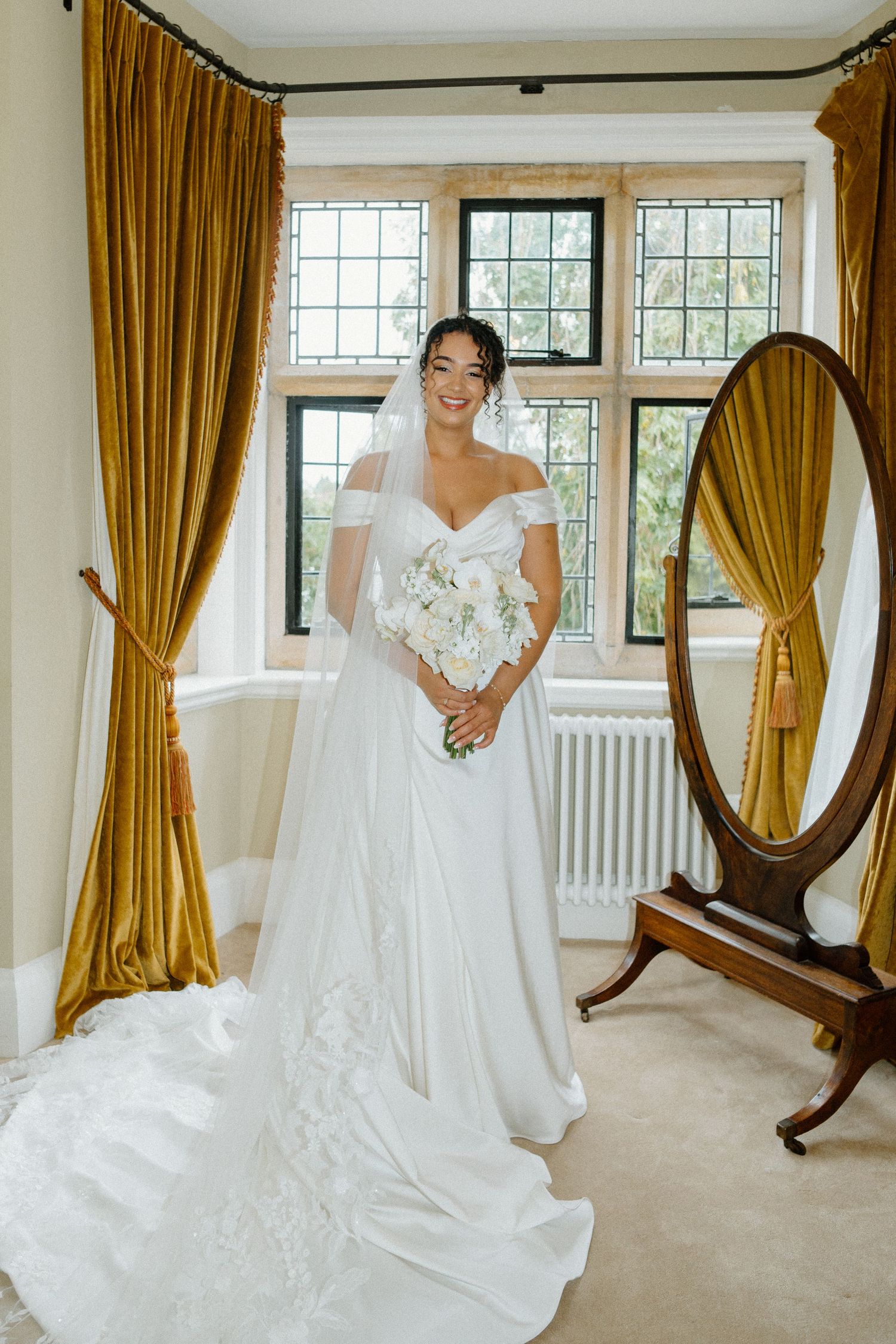 Elegant bride in off-shoulder wedding gown with veil holding white bouquet by a window.