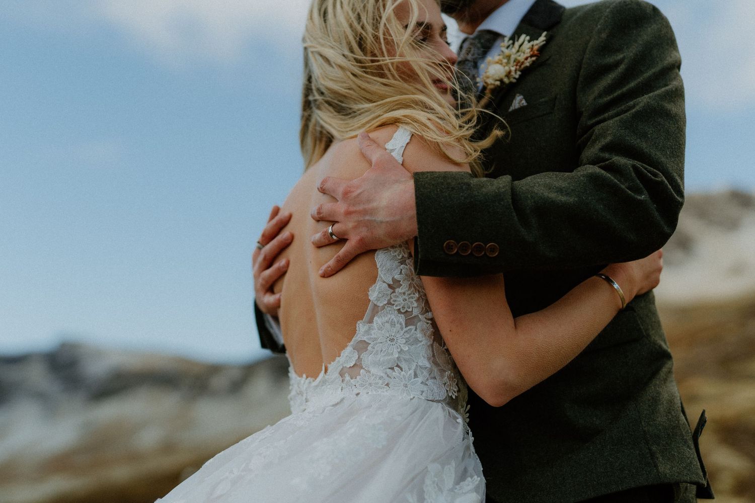 Groom in green tweed suit embraces bride in backless lace wedding gown in mountain landscape.