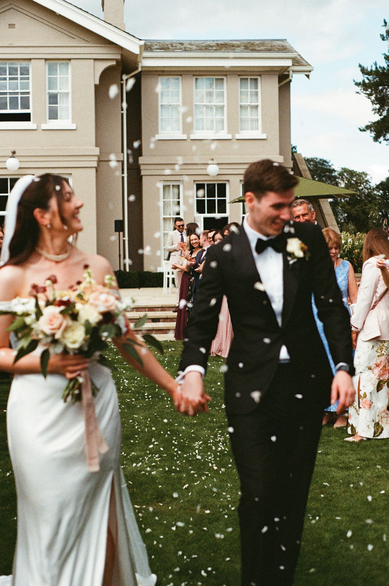 Bride and groom walking through confetti shower on lawn outside elegant country house wedding venue.