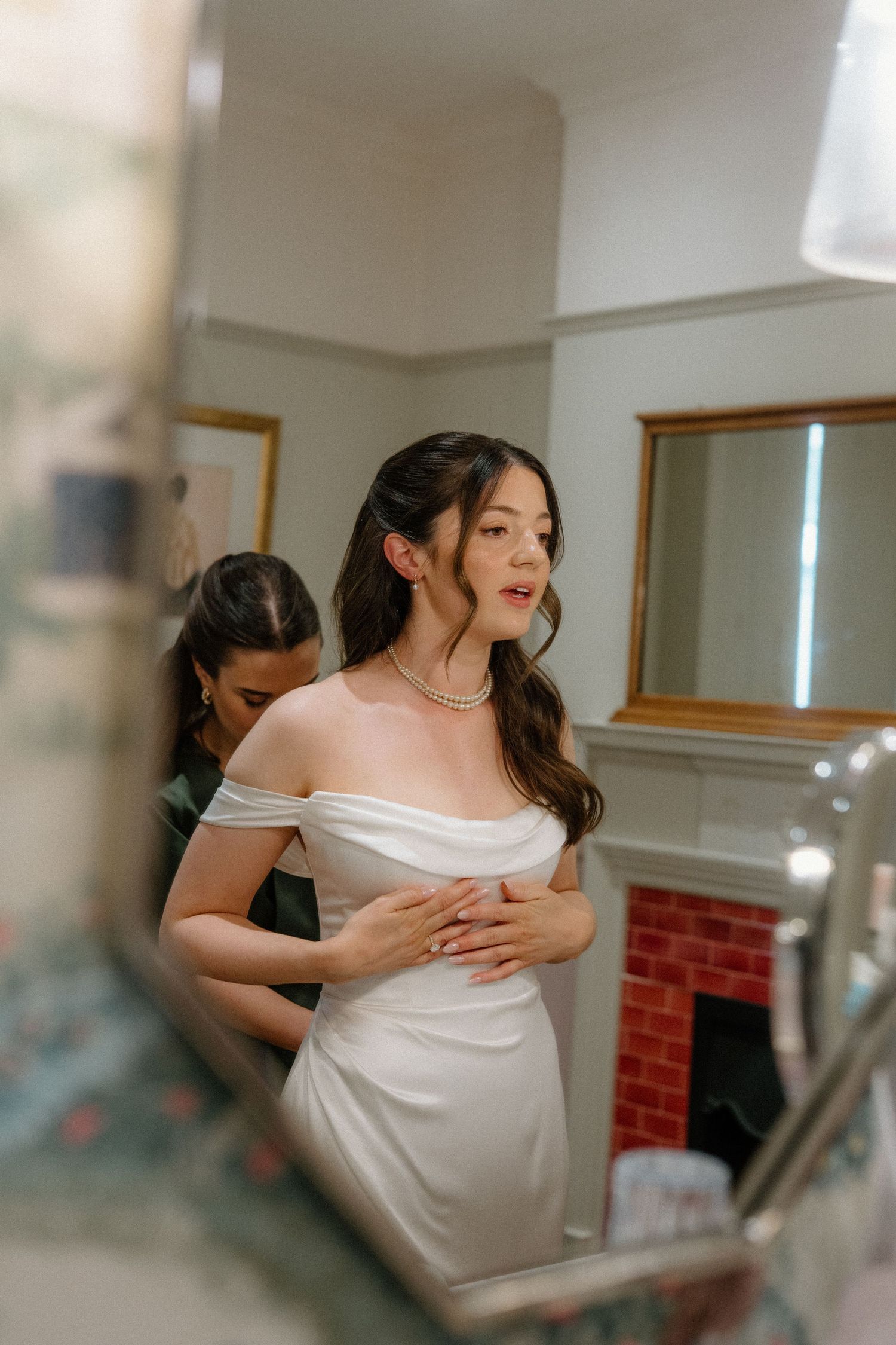 Bride in elegant off-shoulder white wedding dress being helped by bridesmaid in vintage bathroom mirror.