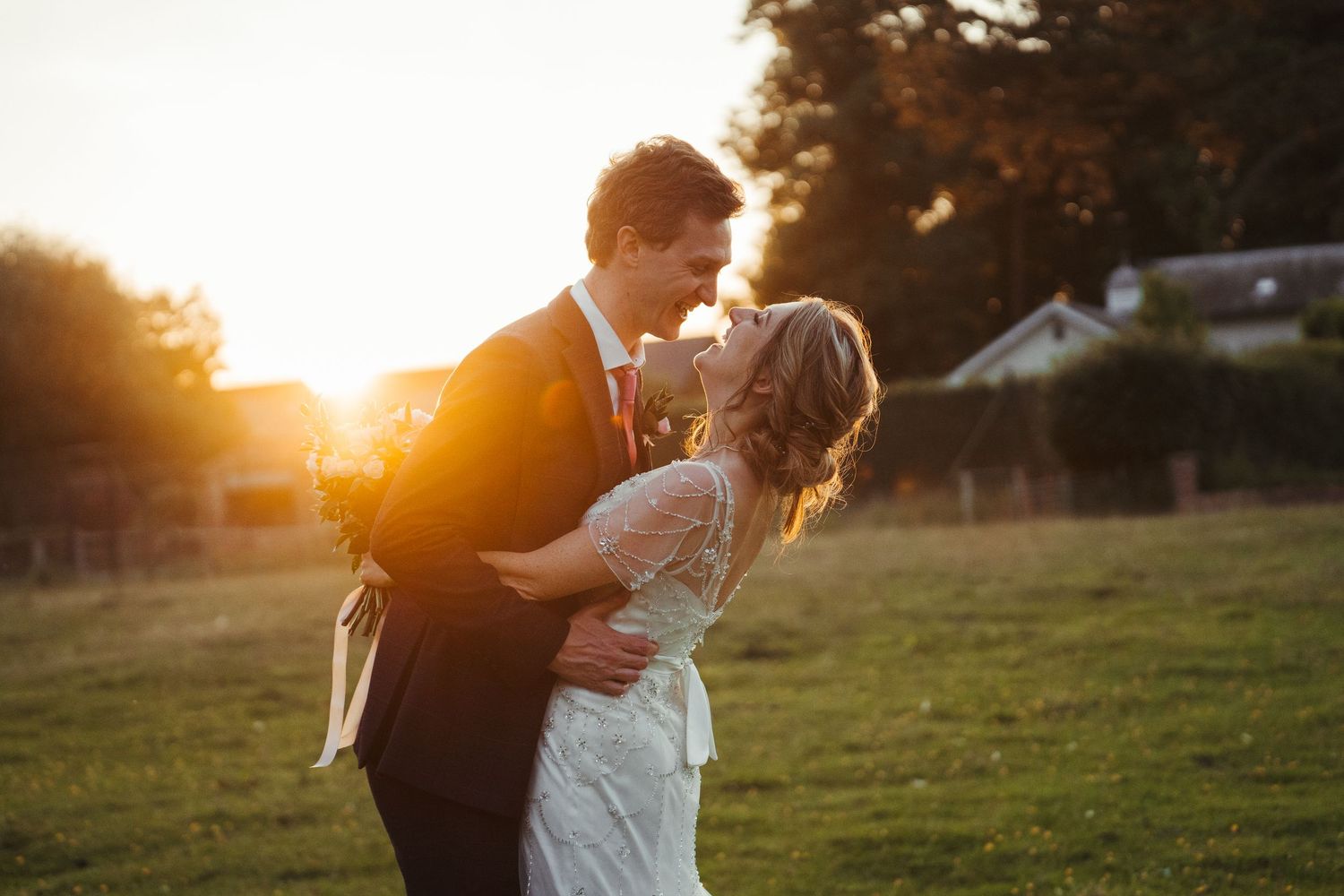 Bride and groom embracing romantically in golden sunset light on lush green wedding day field.