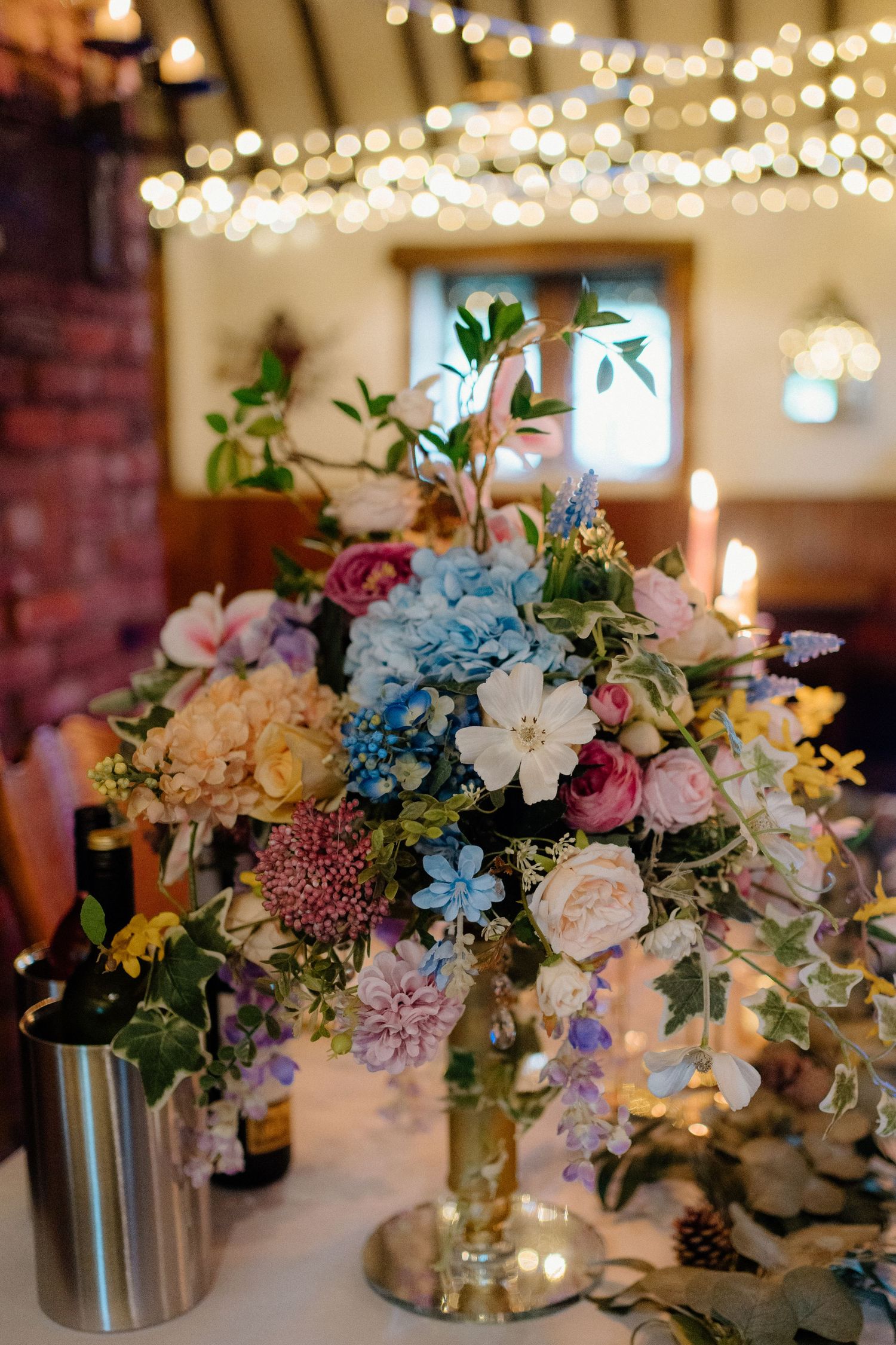 Elegant wedding centerpiece with blue hydrangeas, pink roses and white lilies under twinkling string lights at reception.