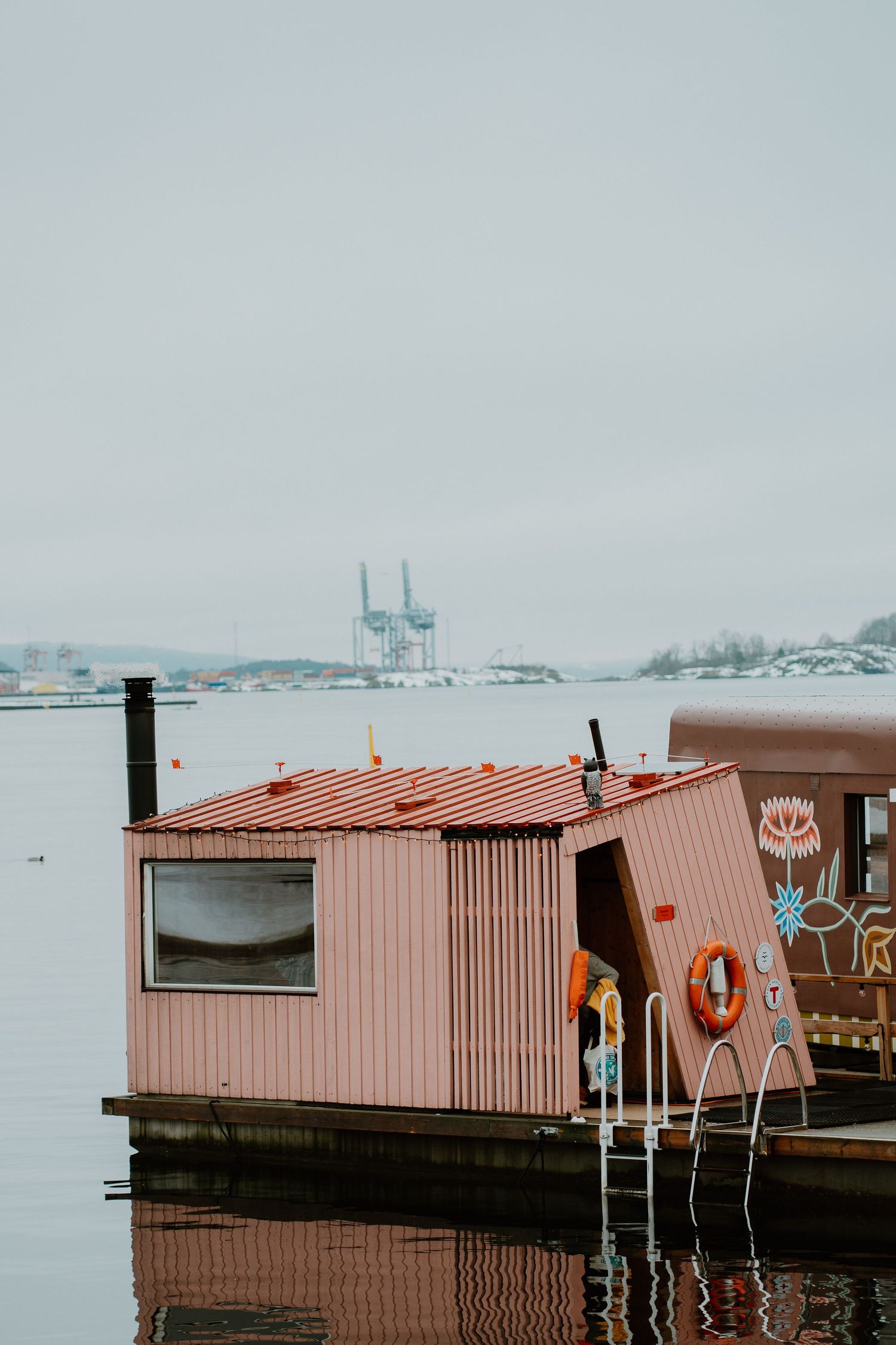 Pink houseboat with a life preserver ring floating on calm water with industrial cranes visible in the background.