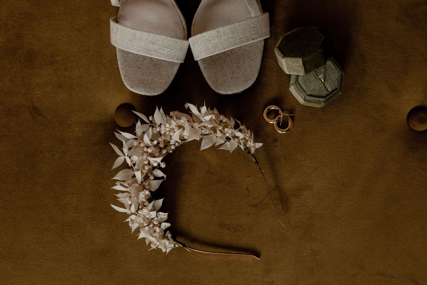 Wedding accessories including white sandals, floral headpiece, and rings arranged on brown velvet.