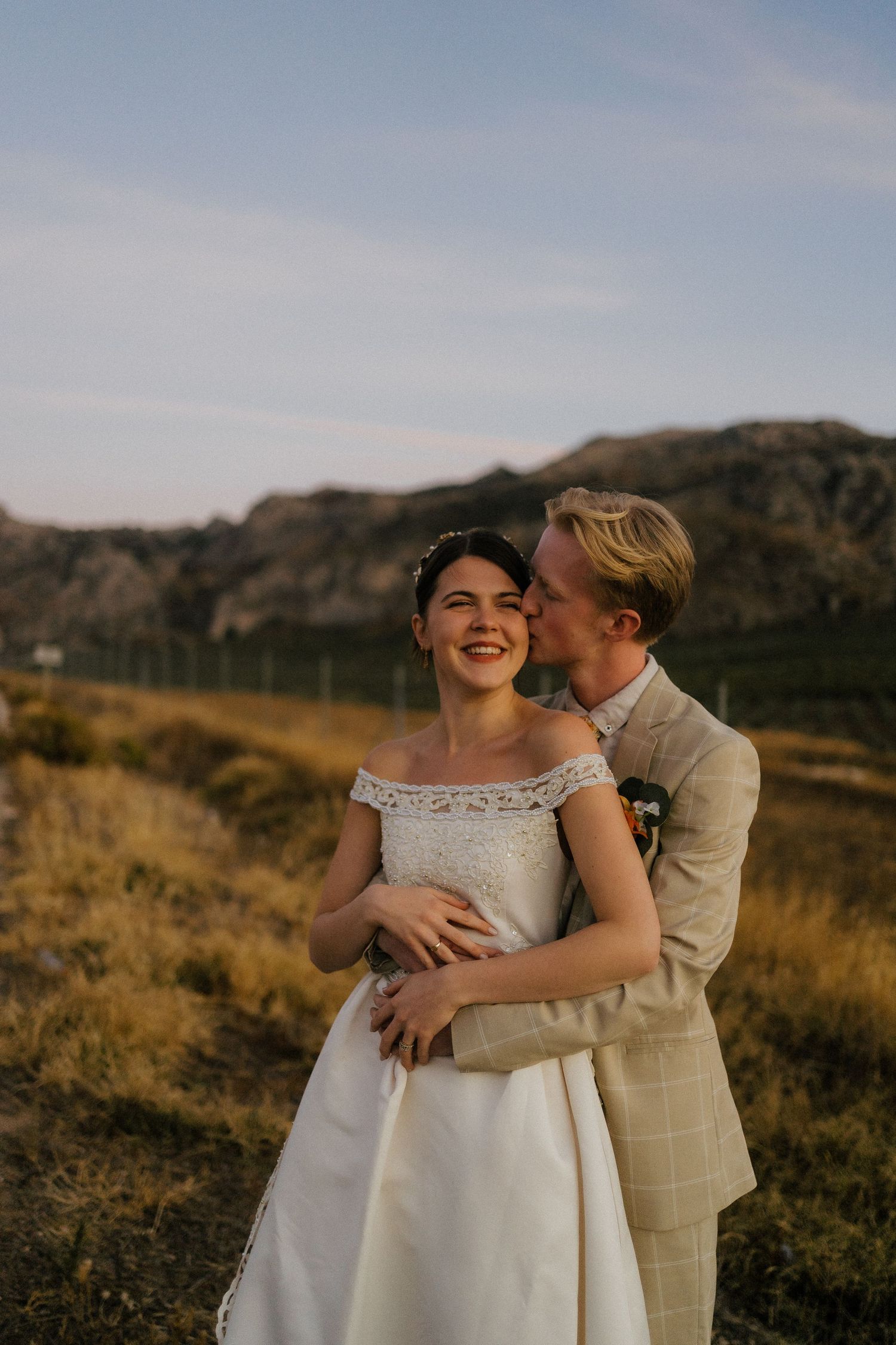 Groom kissing bride on cheek as she smiles joyfully in golden sunlight with mountain scenery.