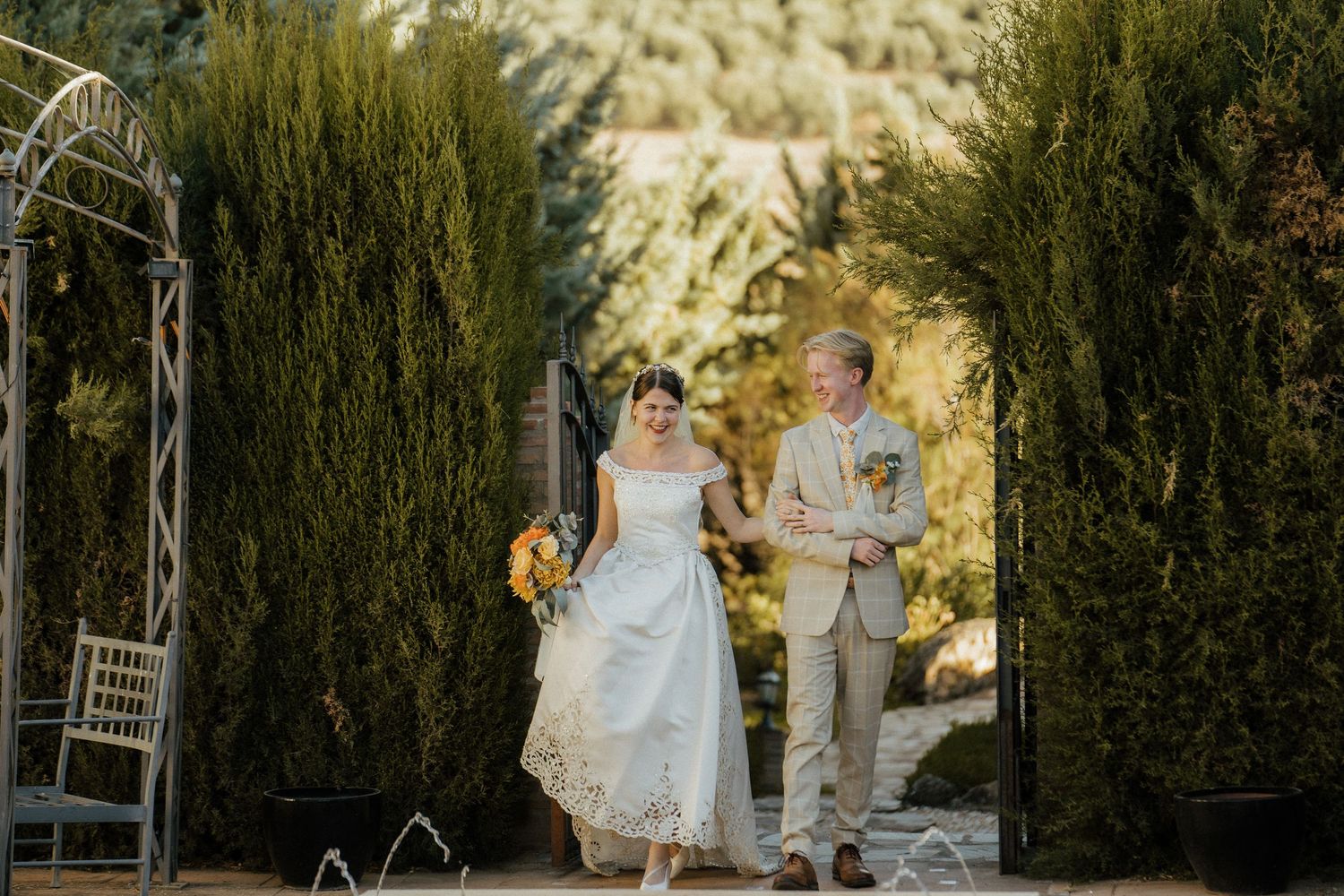 Bride and groom walking hand in hand through cypress tree archway at outdoor wedding.