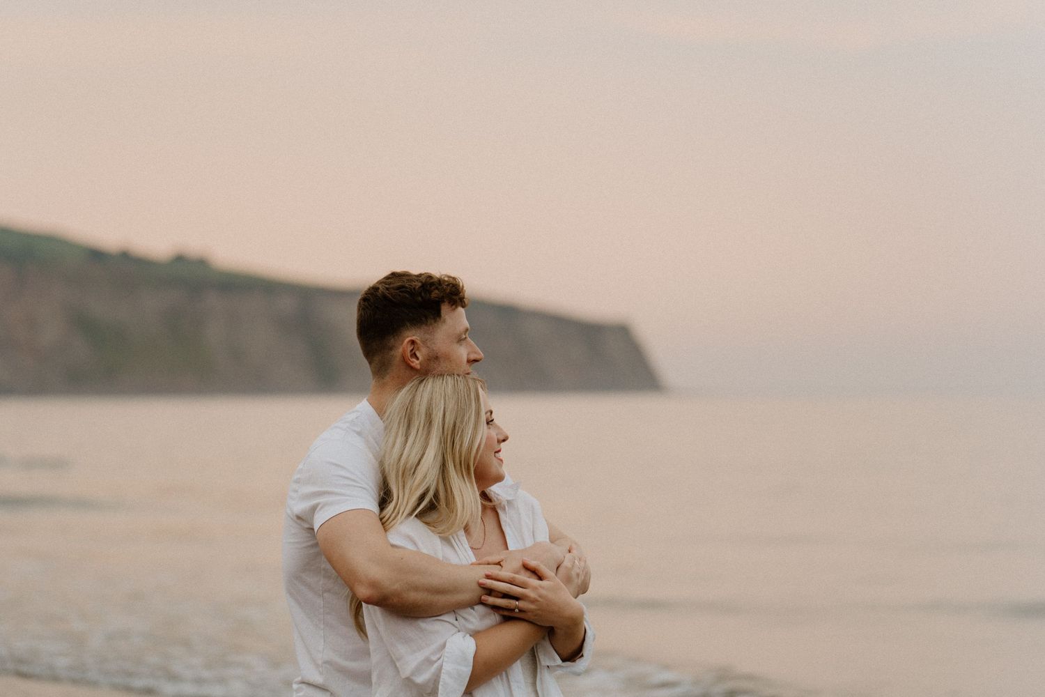 Couple embracing and gazing at the ocean together at sunset near coastal cliffs.