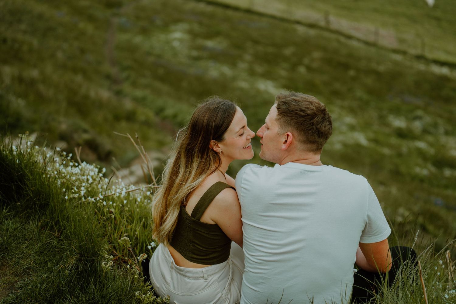 Romantic couple sharing a kiss while sitting together on a green hillside during outdoor couples photography session.