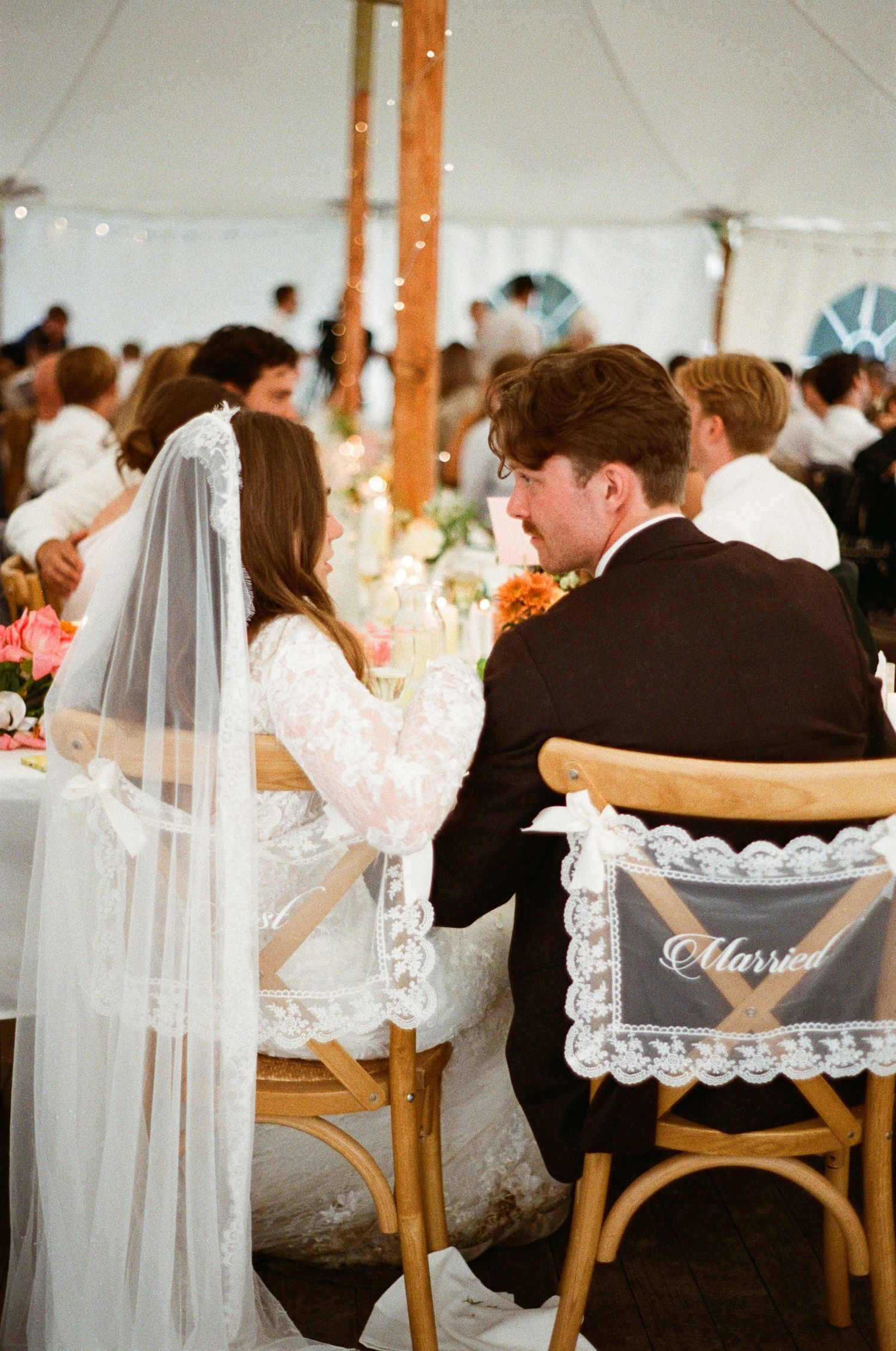Bride and groom seated at wedding reception table under elegant tent, bride wearing lace veil, chairs labeled Married.