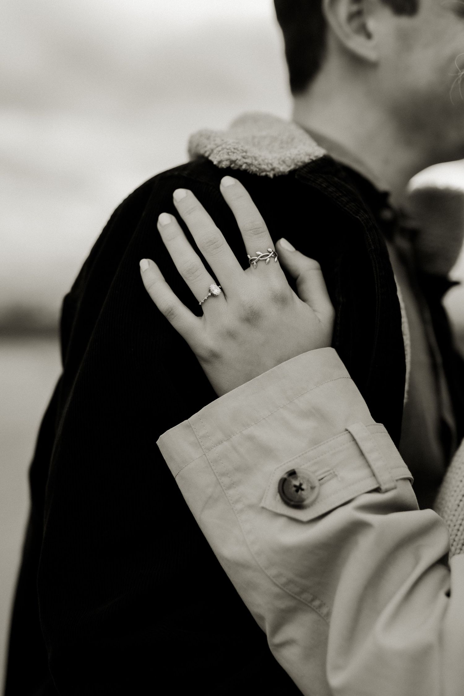 Black and white close-up of womans hand with engagement ring resting on mans black jacket shoulder.