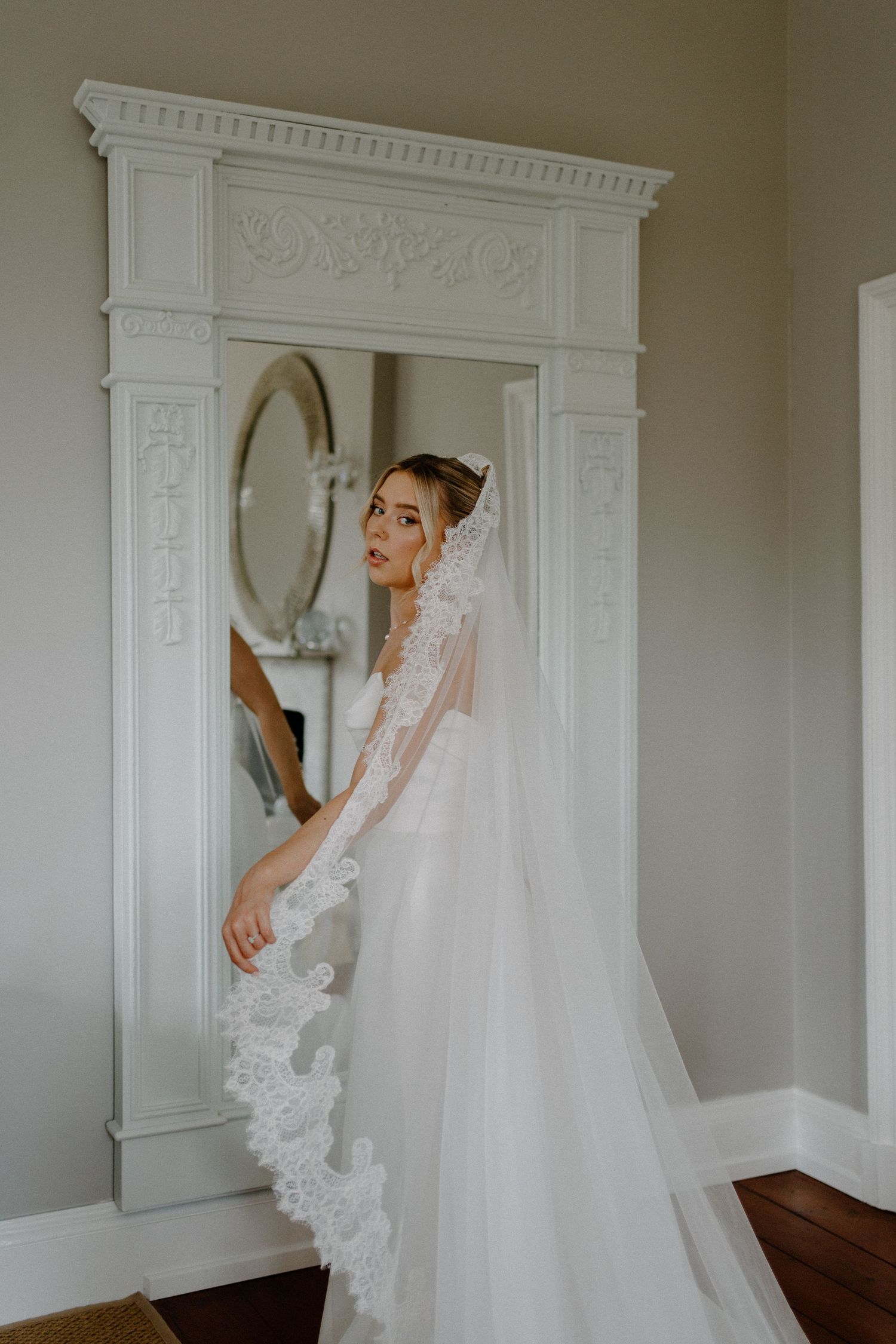 Elegant bride in lace cathedral veil posing beside an ornate white mirror in a classic bridal suite.