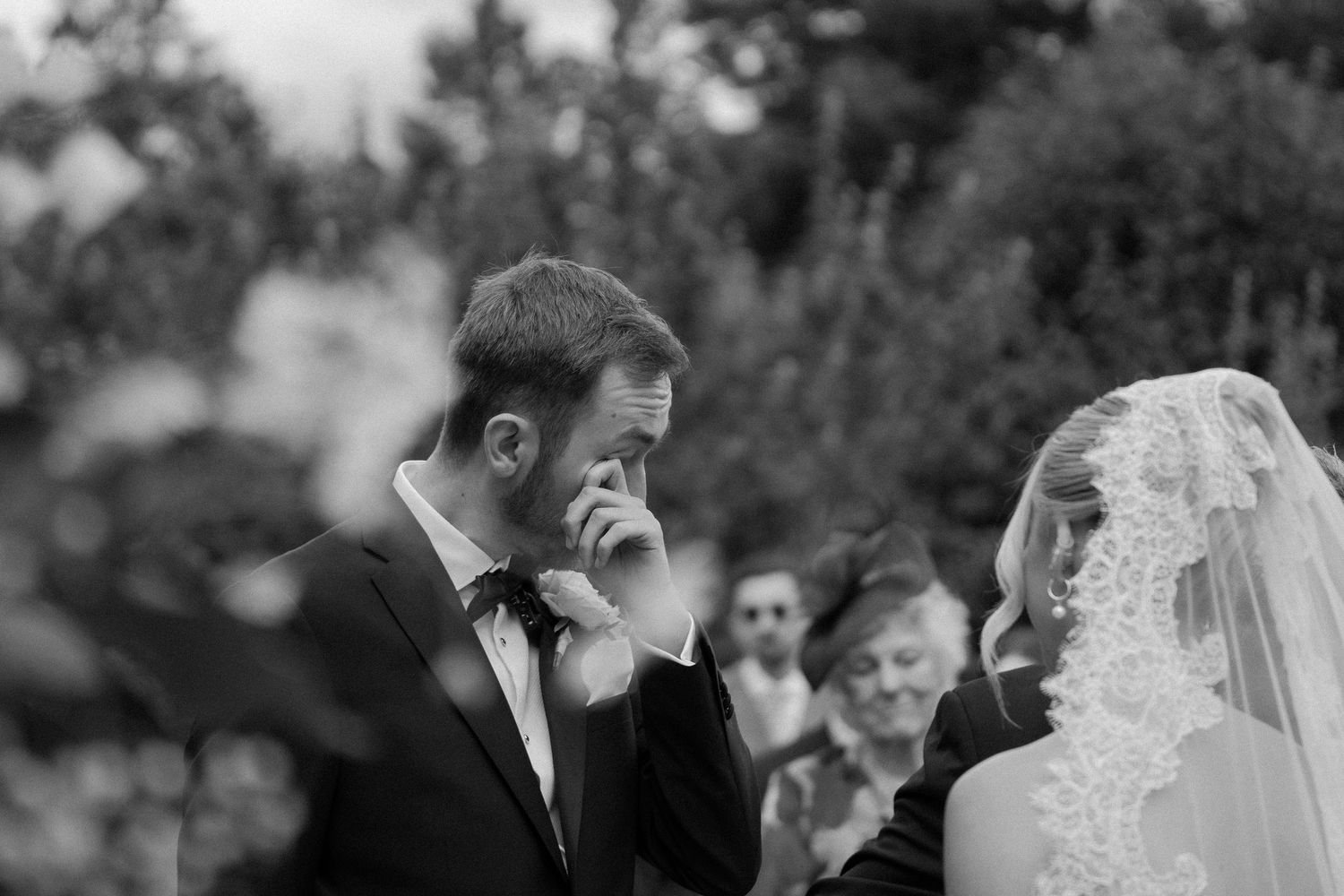 Emotional groom wipes tear during outdoor wedding ceremony beside bride in lace veil, black and white.