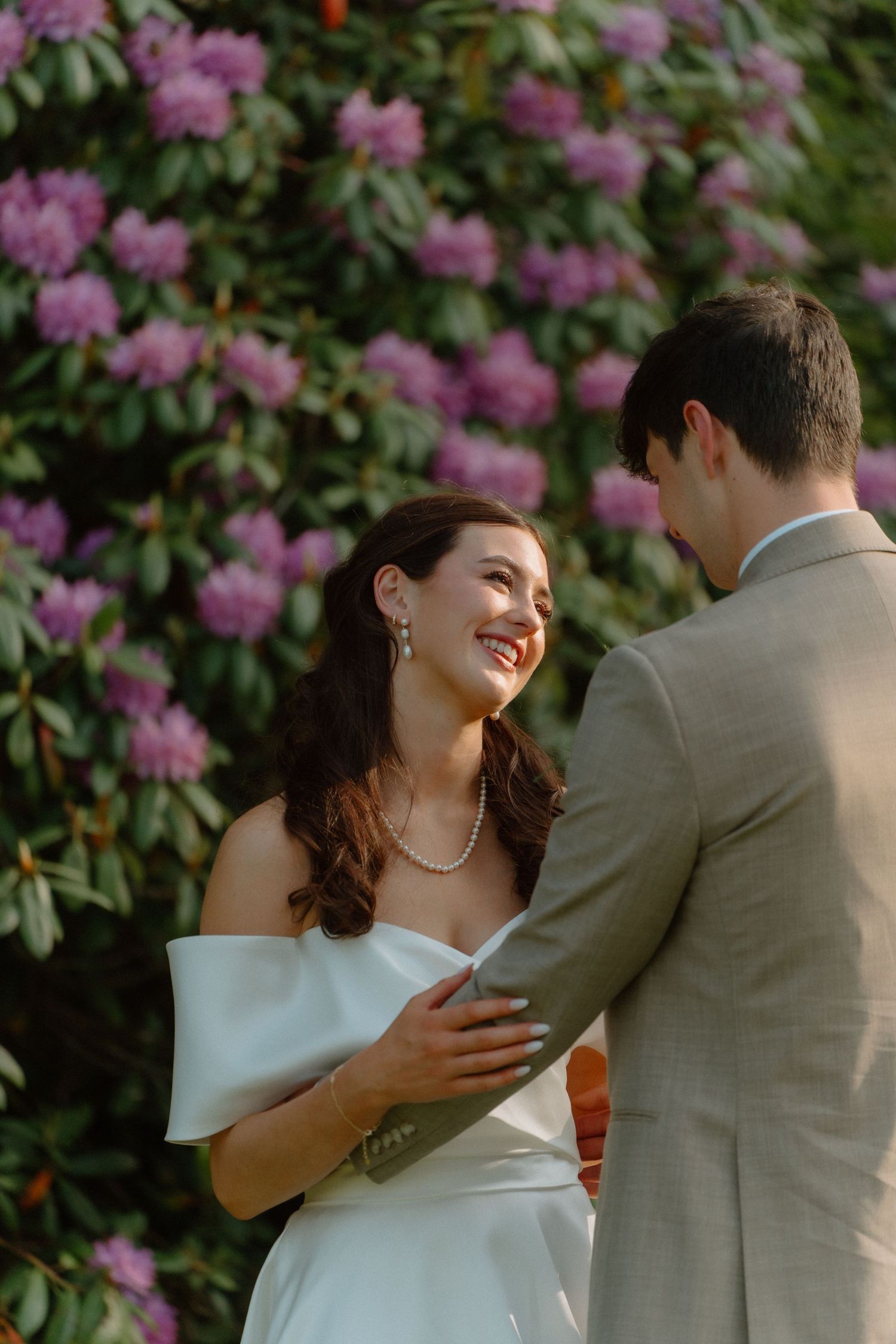 Radiant bride in off-shoulder gown smiling at groom before blooming purple rhododendron bushes.