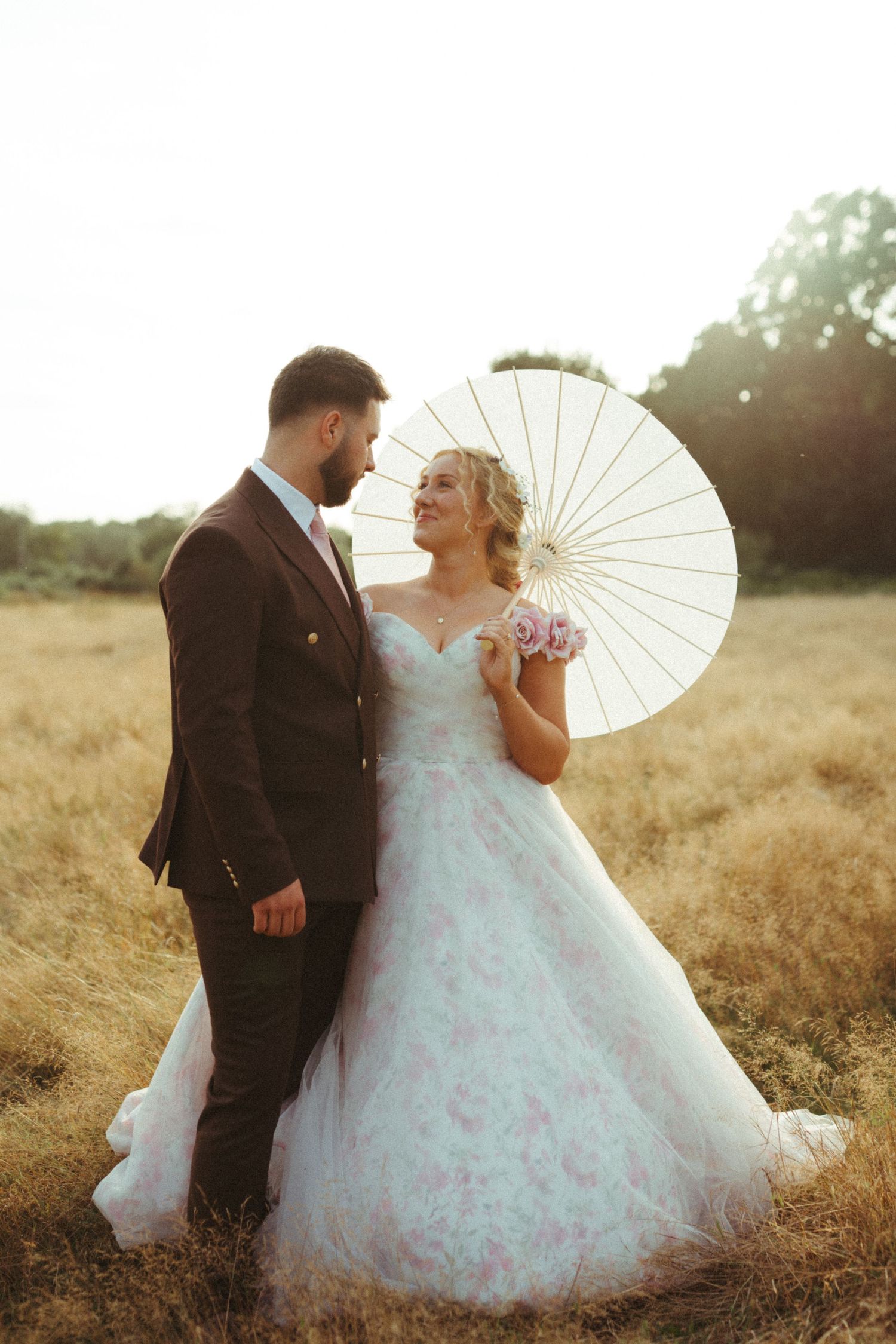 Bride and groom share a romantic moment in sunlit field, bride holding white parasol and pink bouquet.