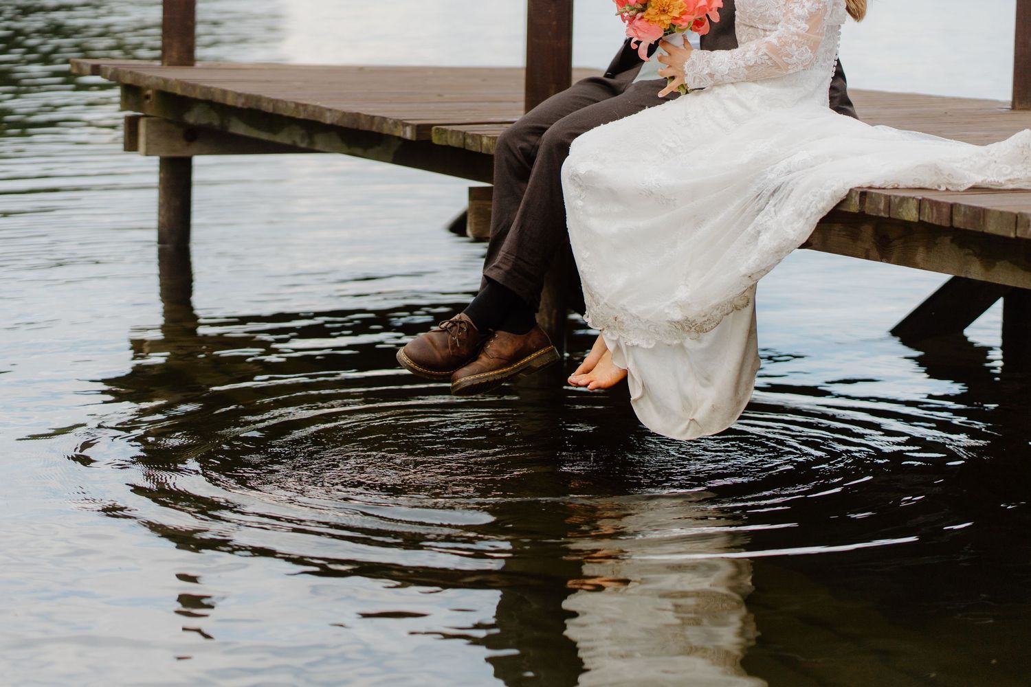 Bride and groom sitting on a wooden dock with feet in the water, bride holding a colorful bouquet.