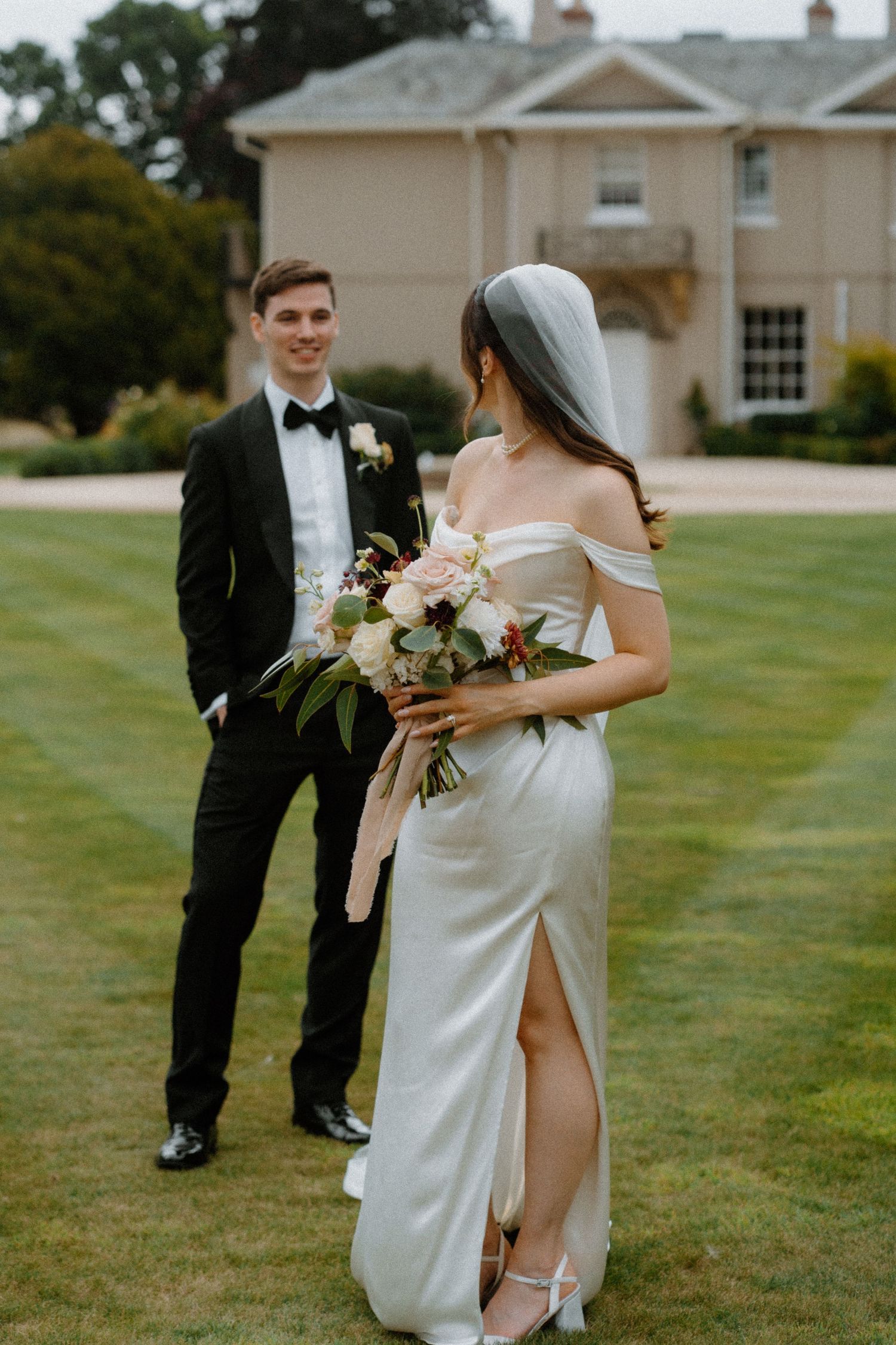 Smiling groom in black tuxedo watches bride in off-shoulder white gown holding bouquet on manicured lawn.