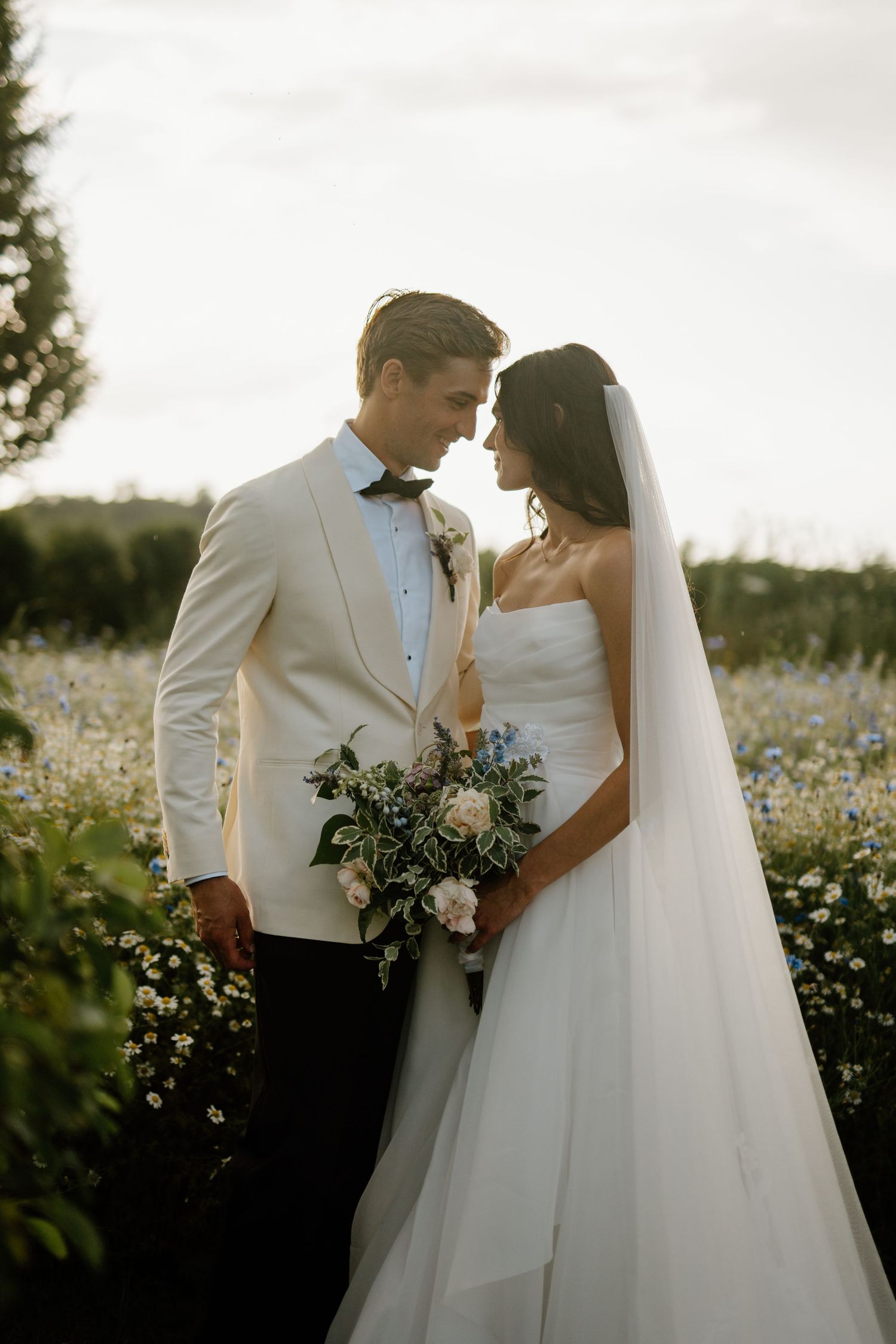 Bride and groom share an intimate moment in a wildflower meadow, groom in ivory tuxedo holding bouquet.