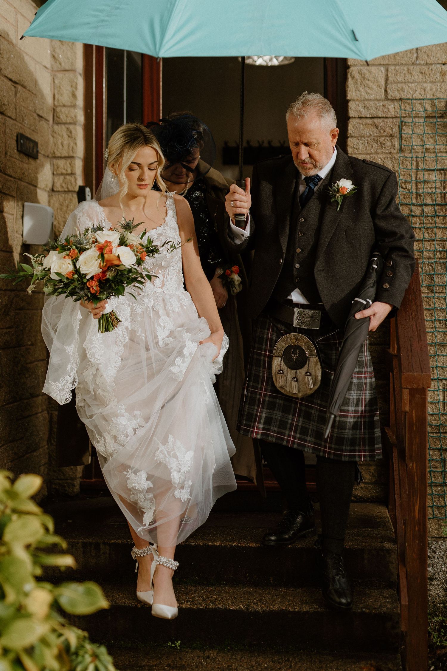 Bride in floral lace dress with bouquet walks out with man in Scottish kilt under teal umbrella.