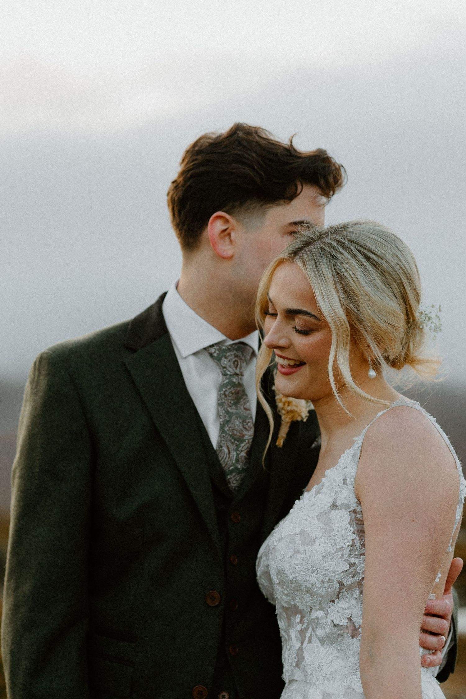 Groom tenderly kisses laughing bride's head in golden light, both dressed in elegant wedding attire outdoors.