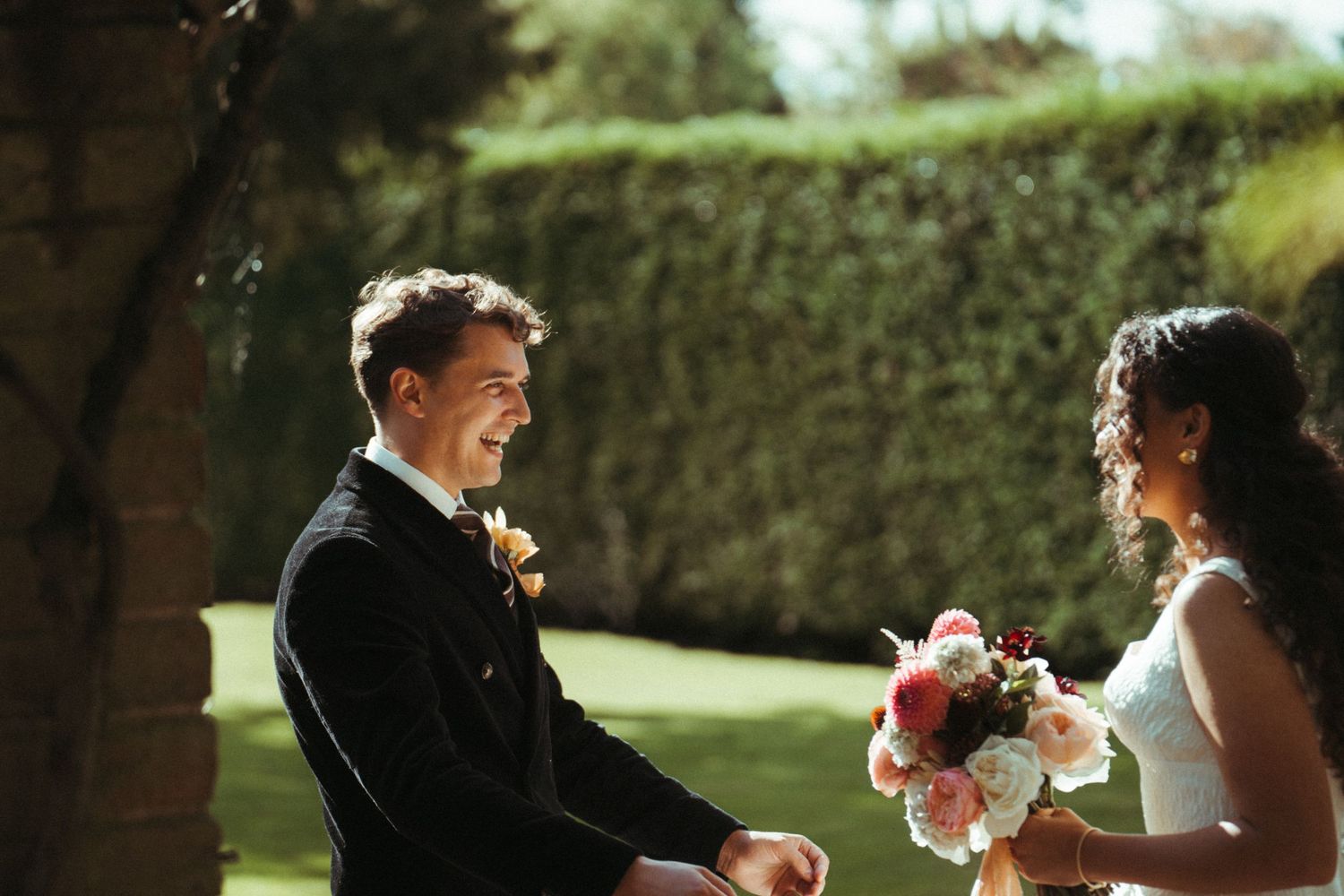 Happy groom in black tuxedo smiling at bride holding pink bouquet during outdoor wedding first look.
