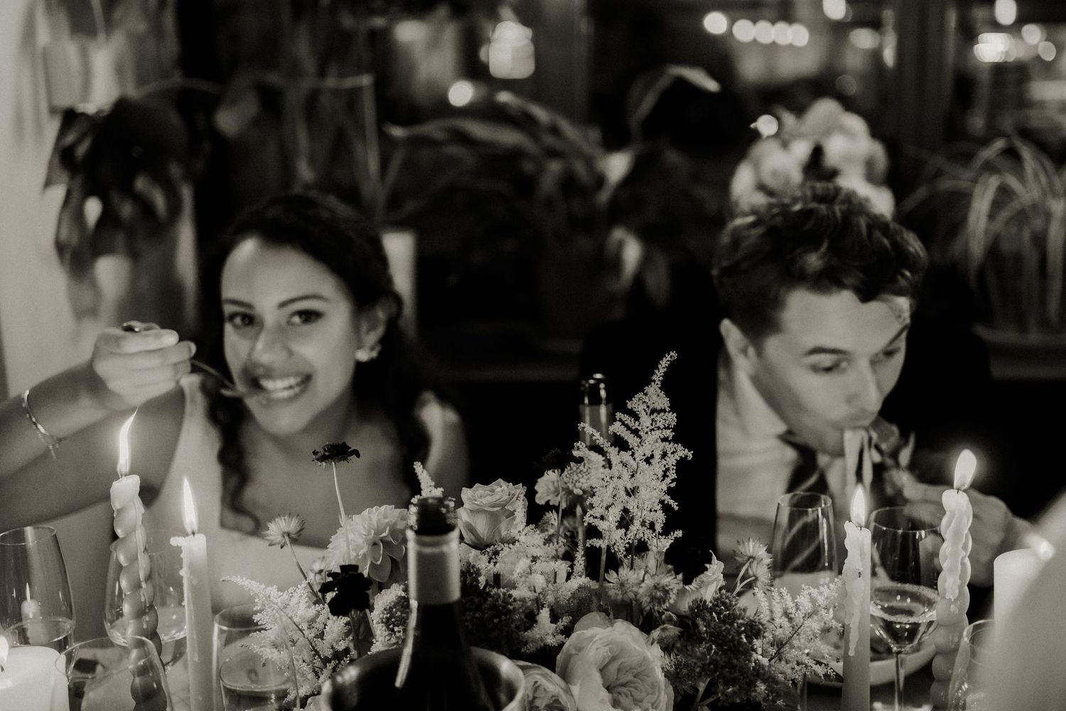 Black and white photo of smiling bride and groom seated at their candlelit wedding reception table.