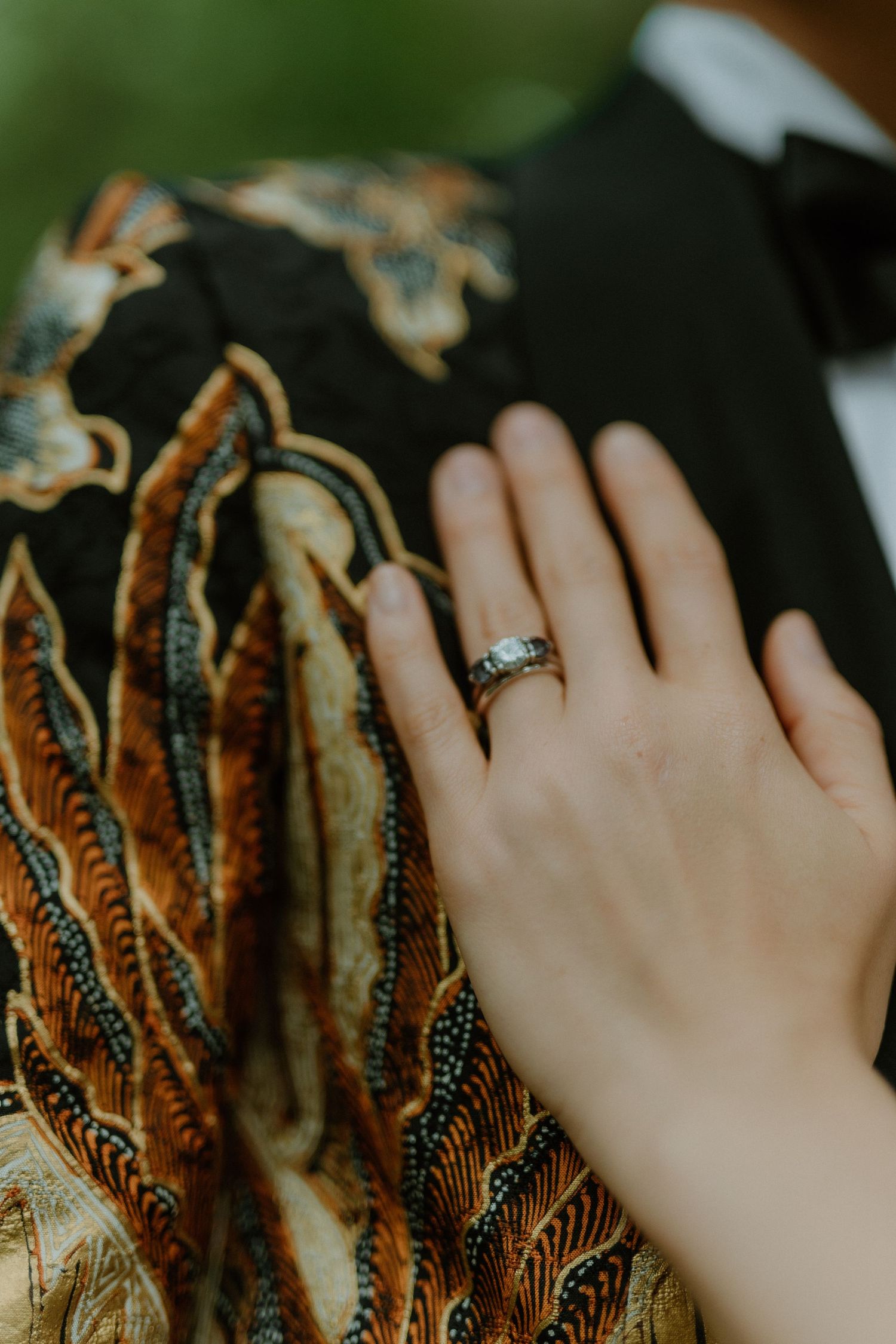 Close-up of bride's hand wearing diamond ring resting on groom's ornate gold batik jacket.