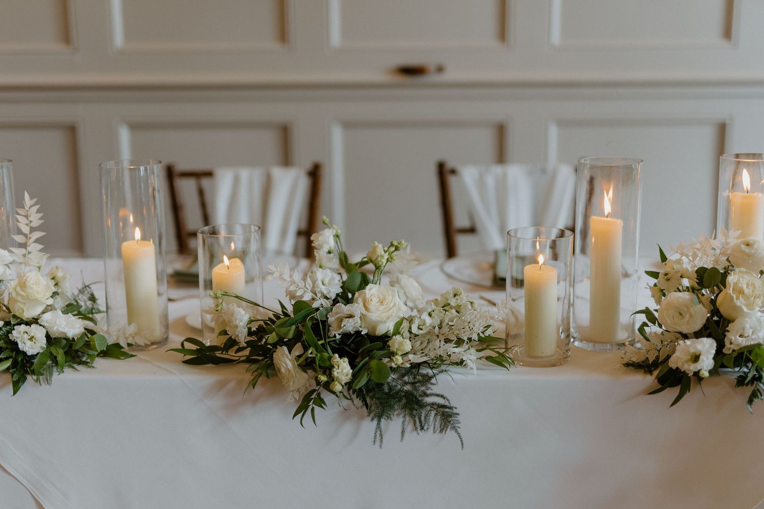 Elegant wedding reception table with white floral centerpiece, greenery, and tall pillar candles.