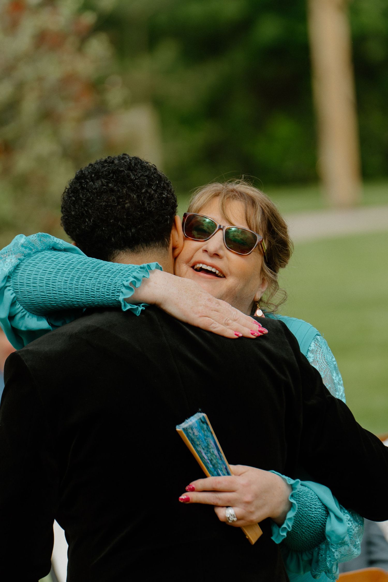A woman in turquoise dress and sunglasses smiles while hugging someone at an outdoor wedding reception.