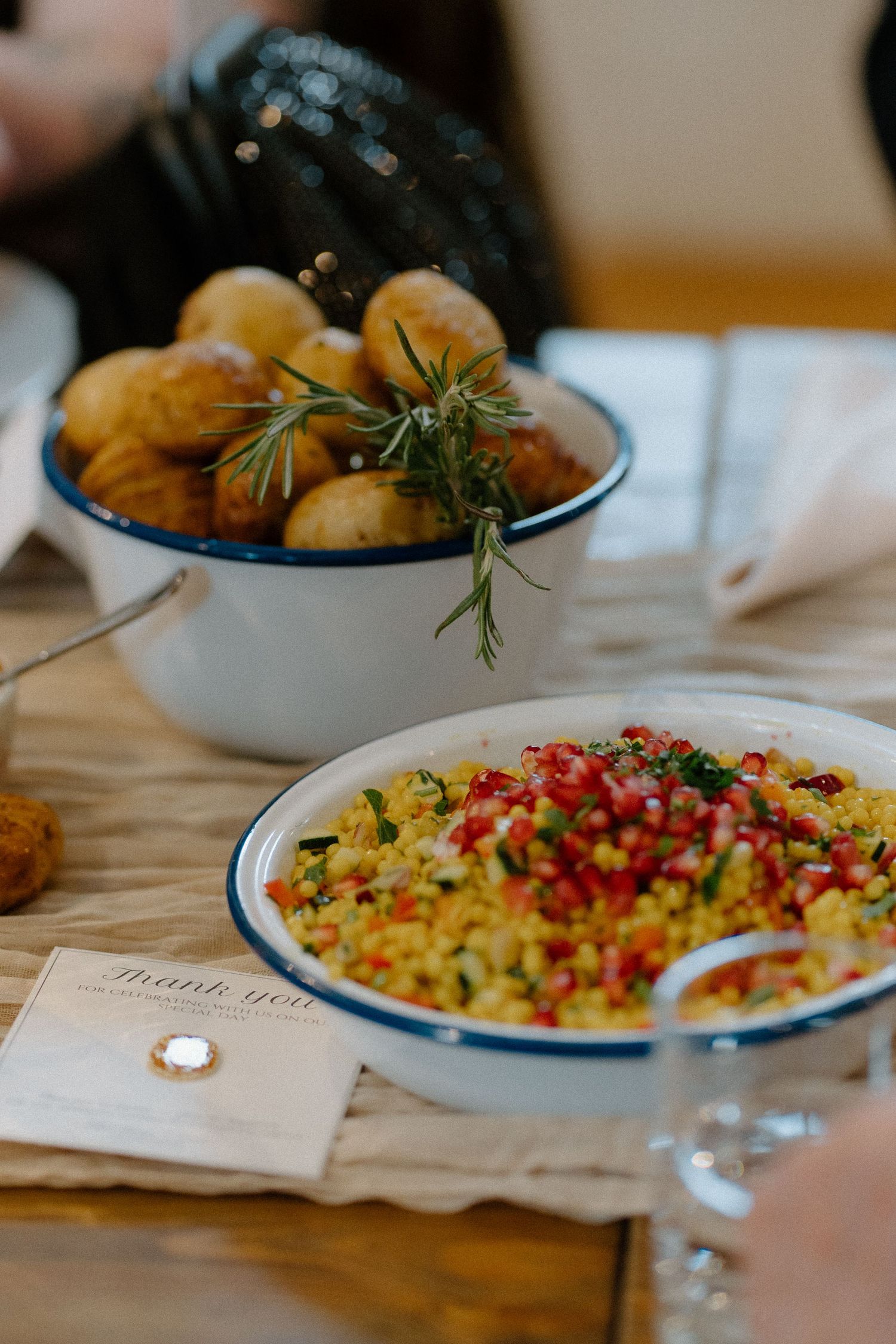 Close-up of rustic dinner table with bowl of potatoes and herbs beside colorful rice dish garnished with pomegranate seeds.