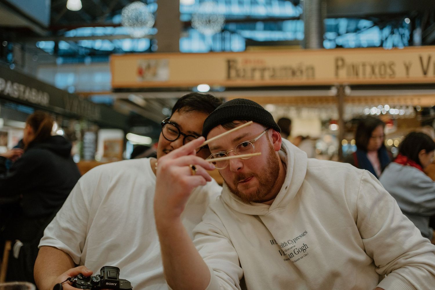 Two friends sit together at a restaurant table, with one making a playful gesture while wearing glasses.