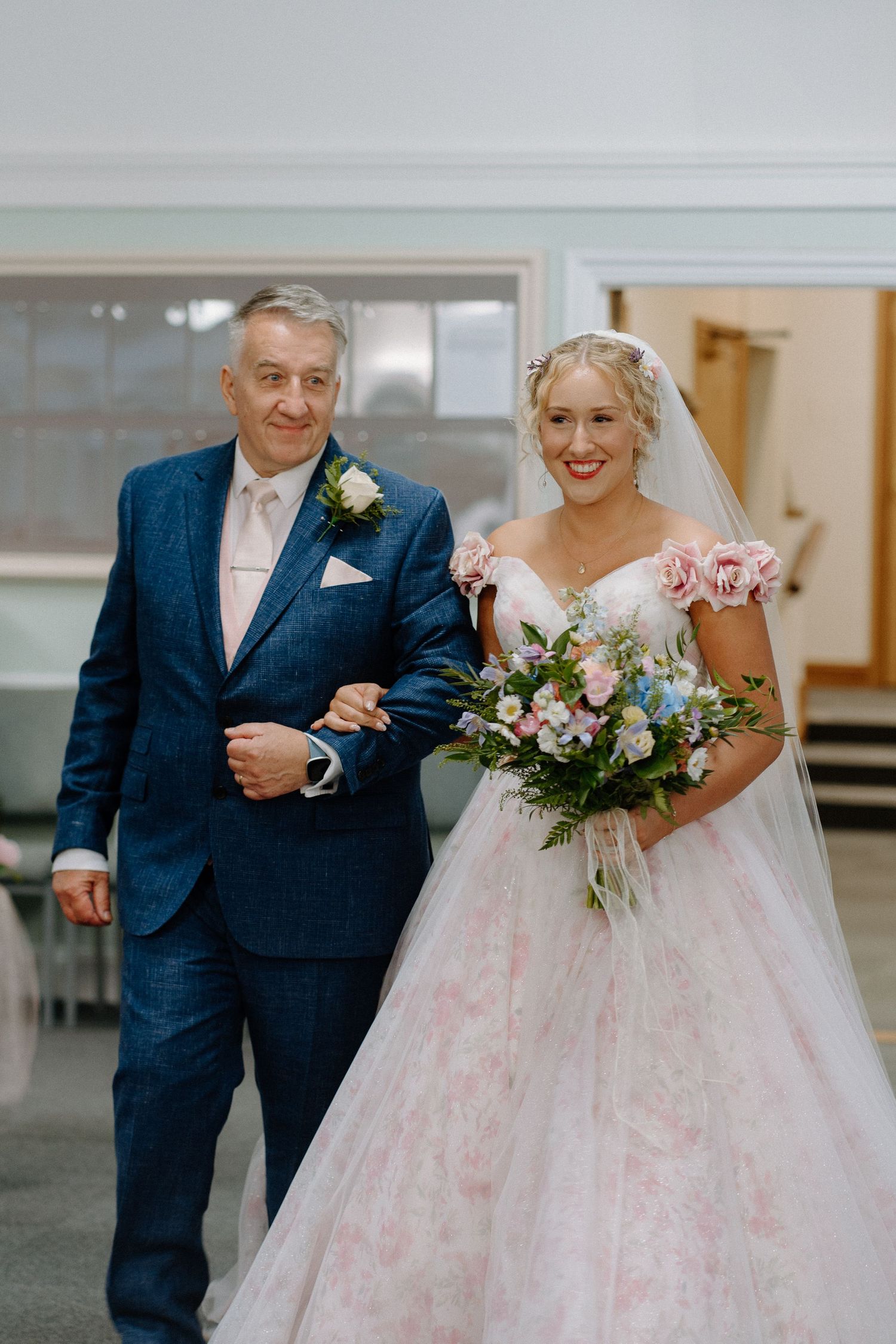 A couple walking arm in arm at their wedding, with the bride in a pink-tinted gown holding a pastel floral bouquet.