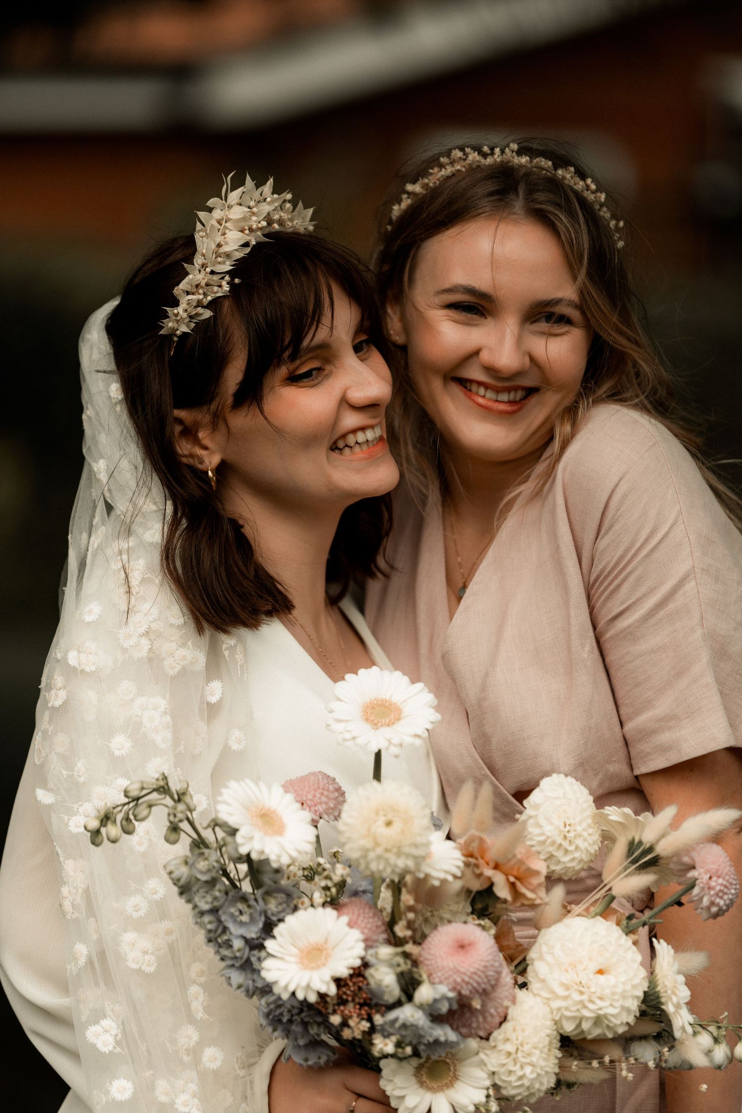Two wedding attendees pose with bright smiles and a stunning floral bouquet outdoors.