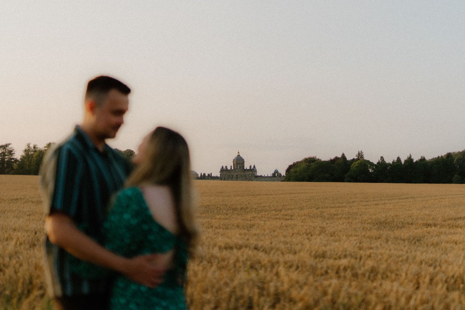 A couple embraces in a golden wheat field at sunset, with forest silhouettes on the horizon.