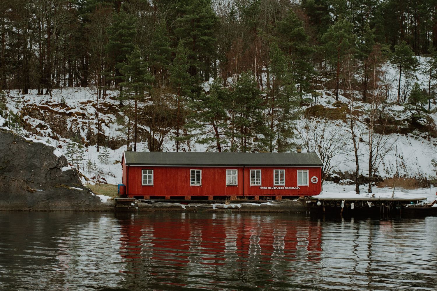 Red floating house reflecting in calm waters against snowy Norwegian landscape.