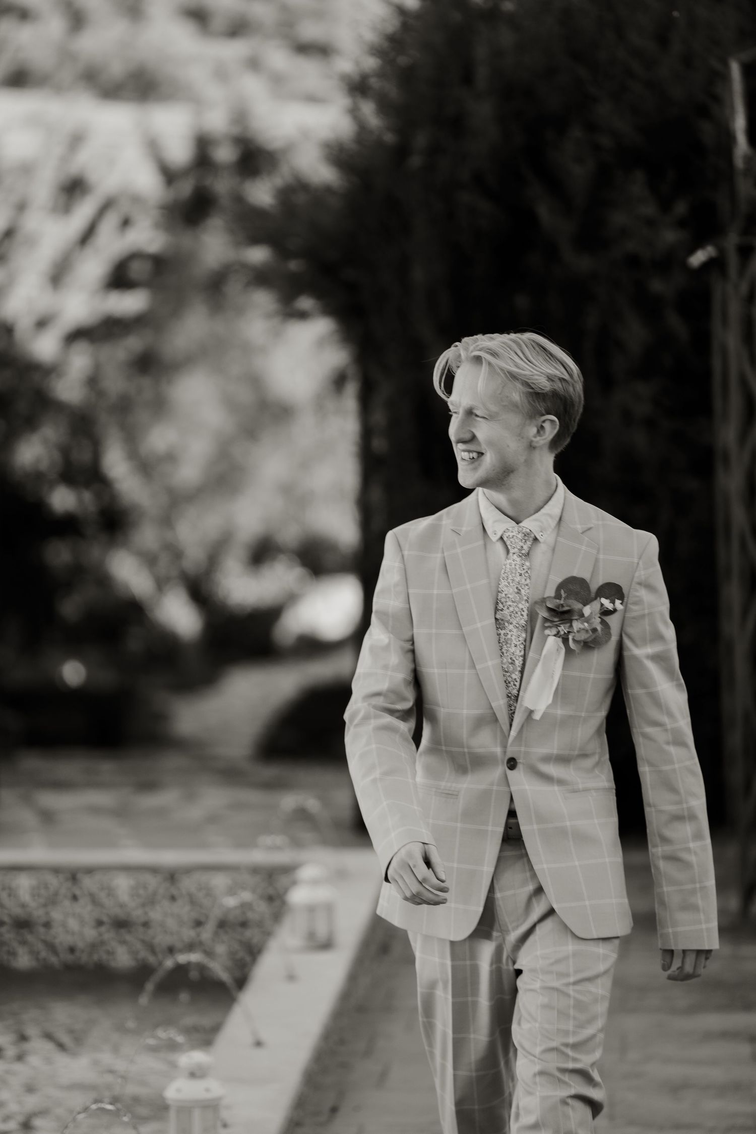 Black and white portrait of smiling groom in plaid suit with boutonniere at outdoor wedding.