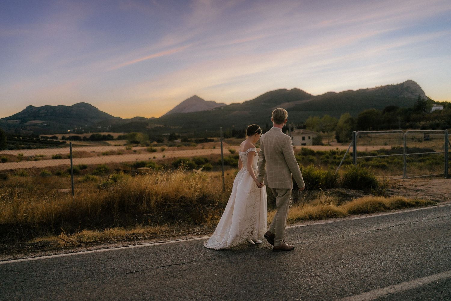 Bride and groom holding hands walking down a rural road at sunset with mountain backdrop.