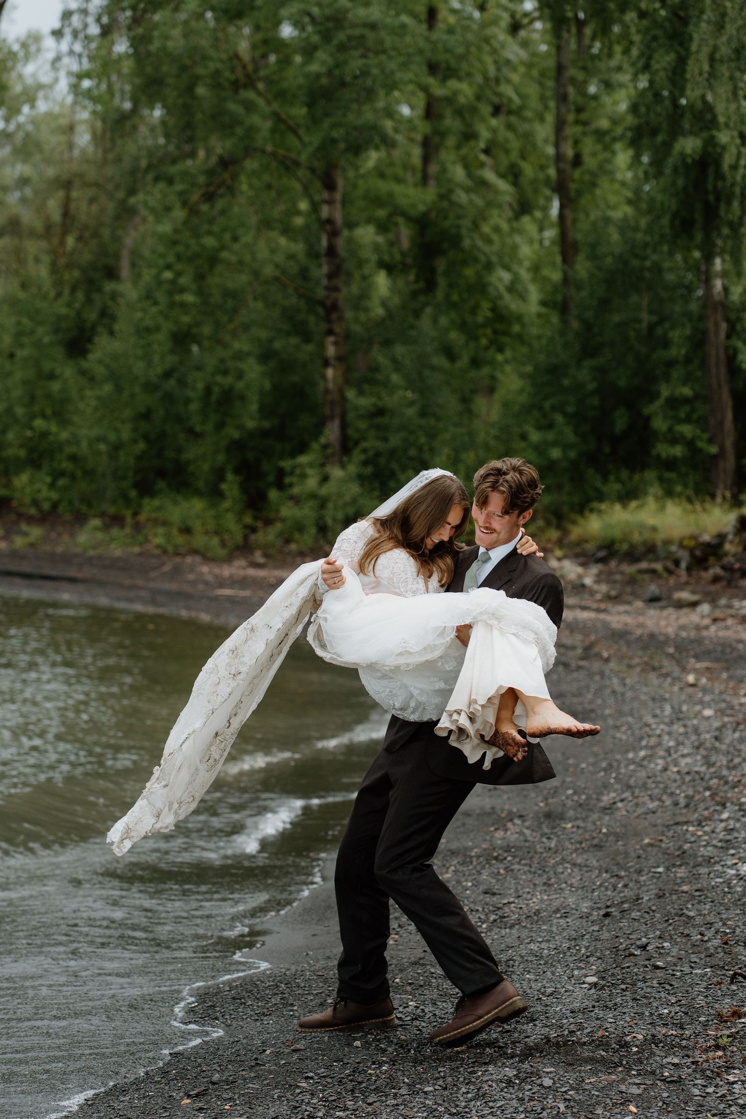 Groom carries barefoot bride in lace gown along a rocky lakeside shore among green trees.