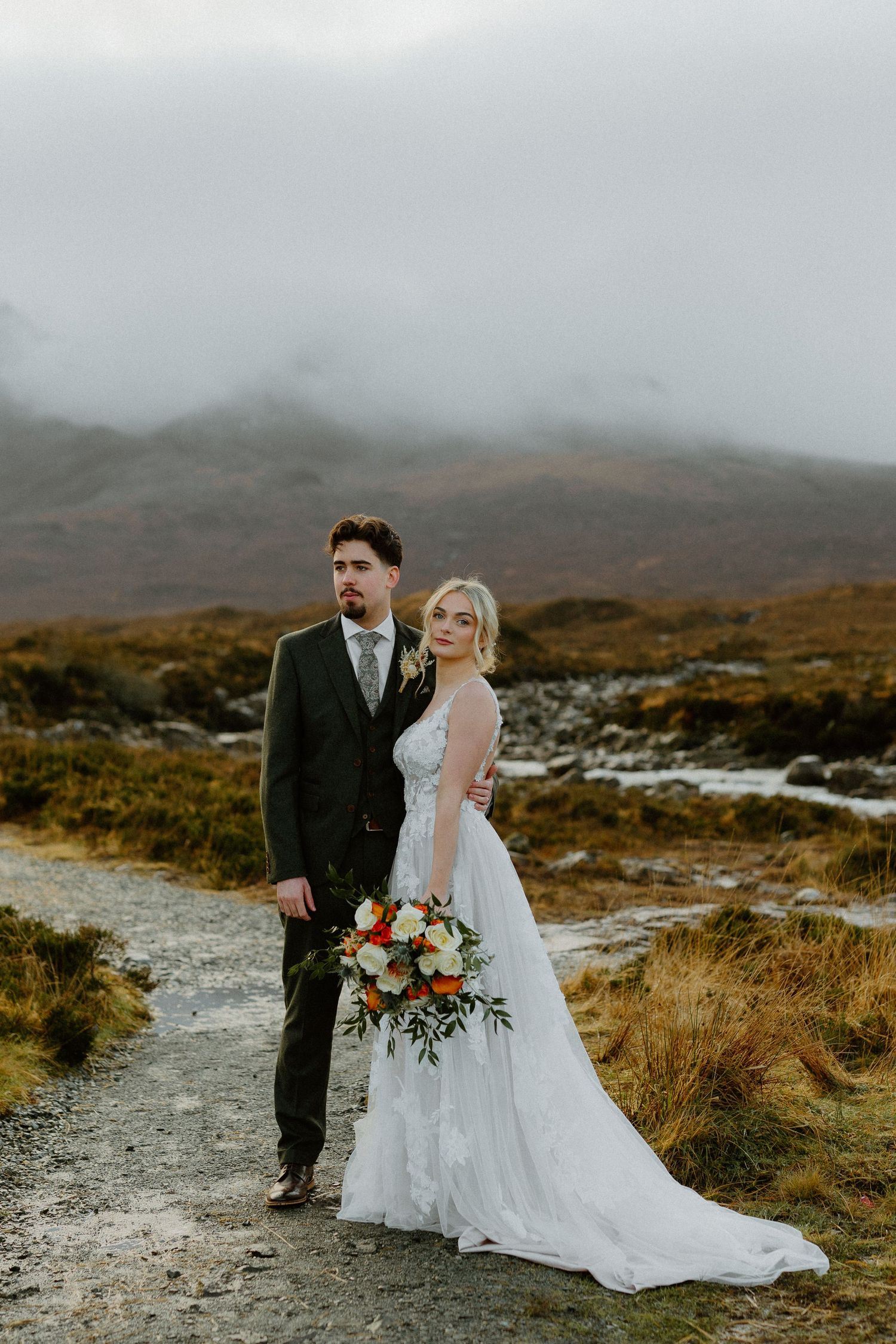 Bride holding a colorful bouquet and groom in dark suit pose on a misty Highland path.