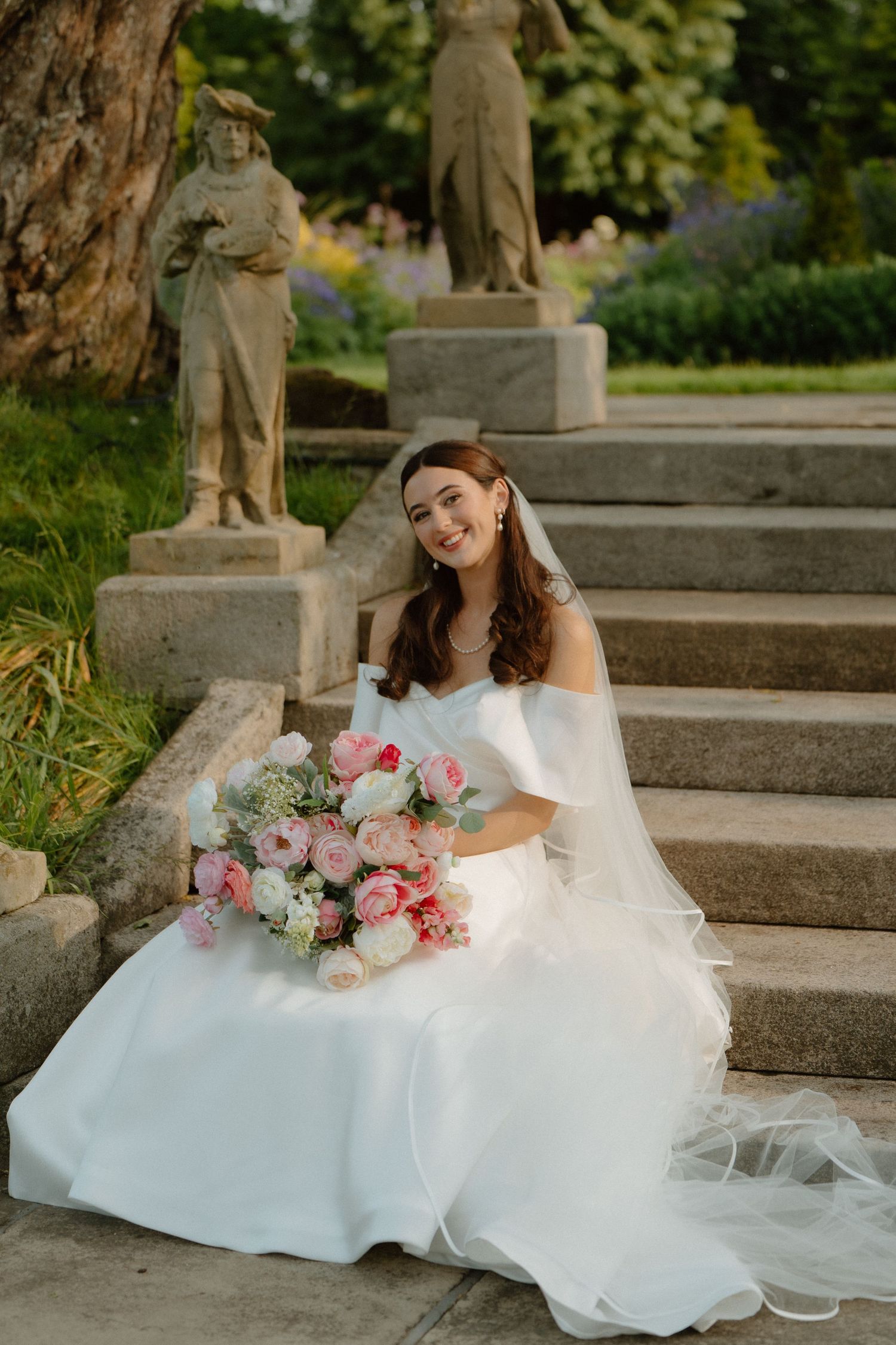 Elegant bride in white gown sitting on stone steps with pink peony bouquet and veil in garden.