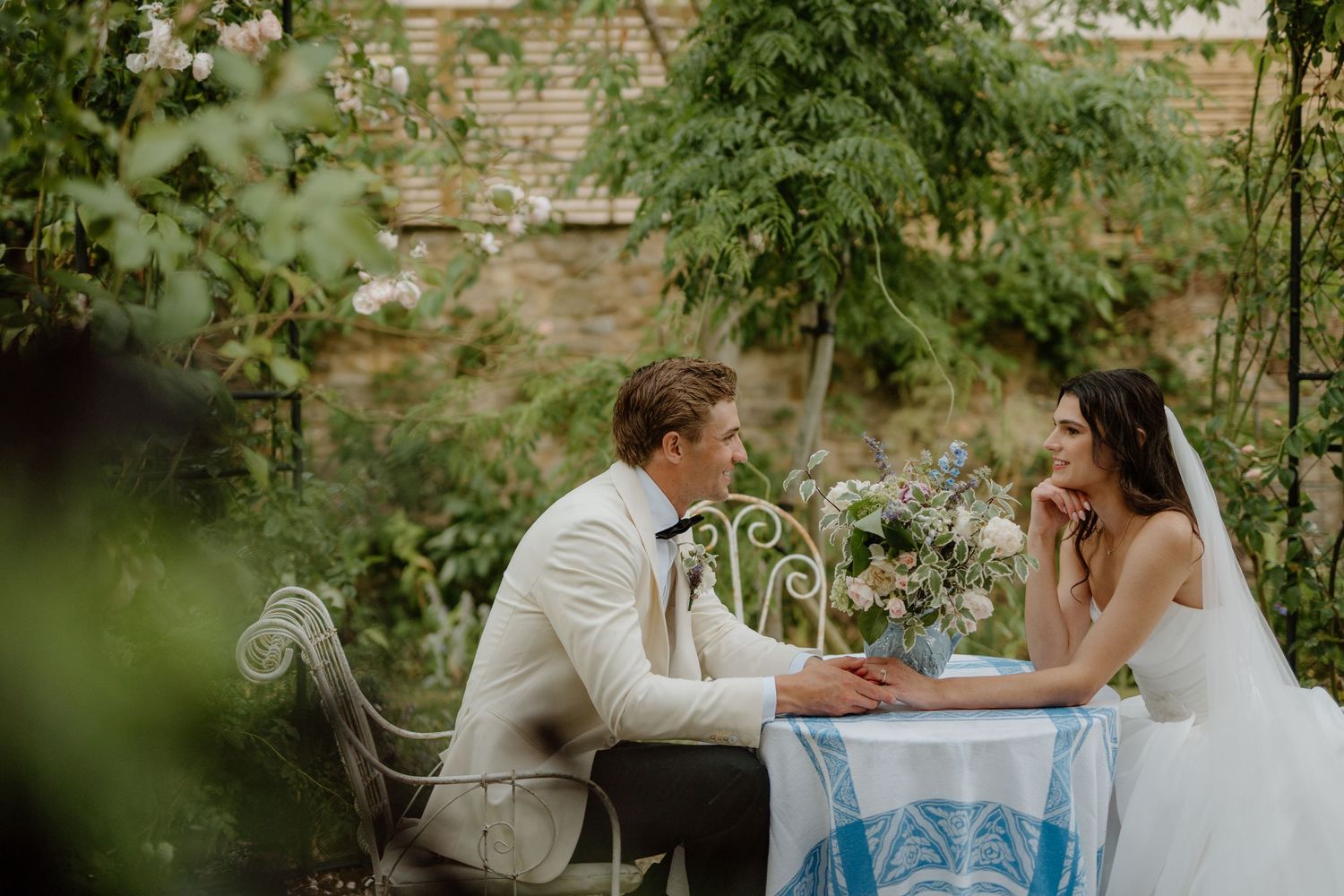 Bride and groom sit together at an elegant outdoor garden table adorned with a blue floral centerpiece.