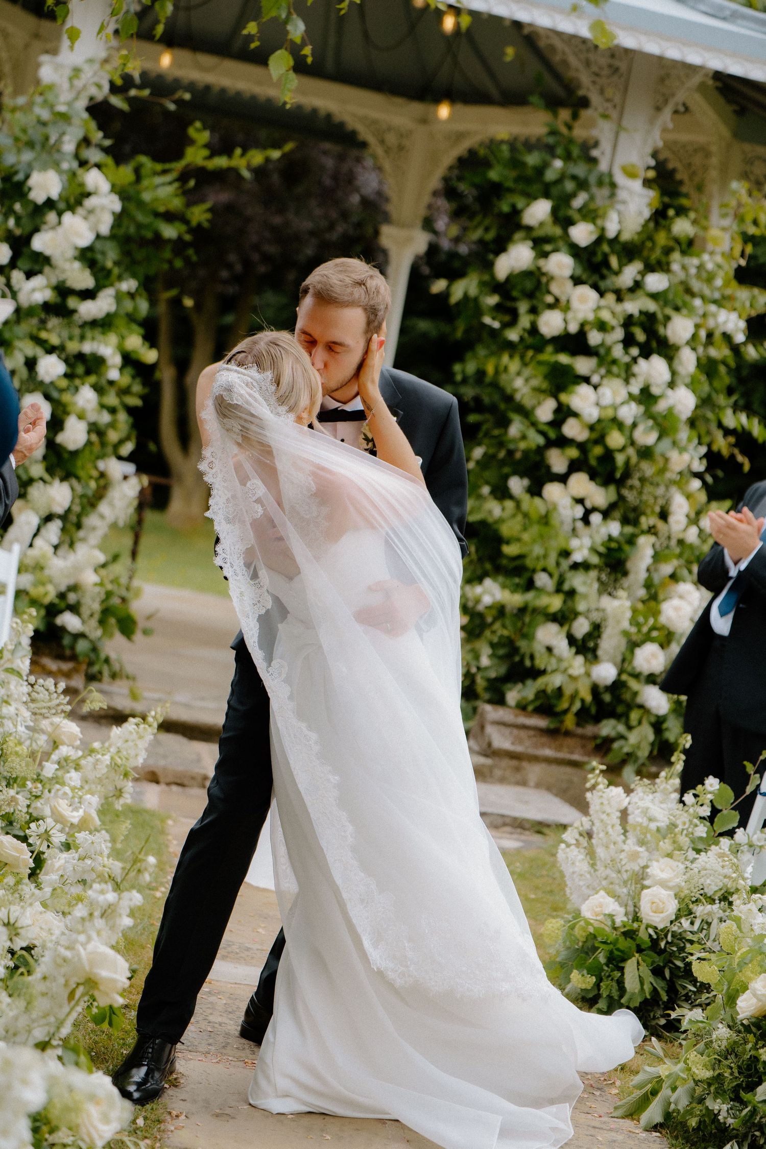 Groom embraces bride for first kiss under white floral pergola arch at elegant outdoor garden wedding.