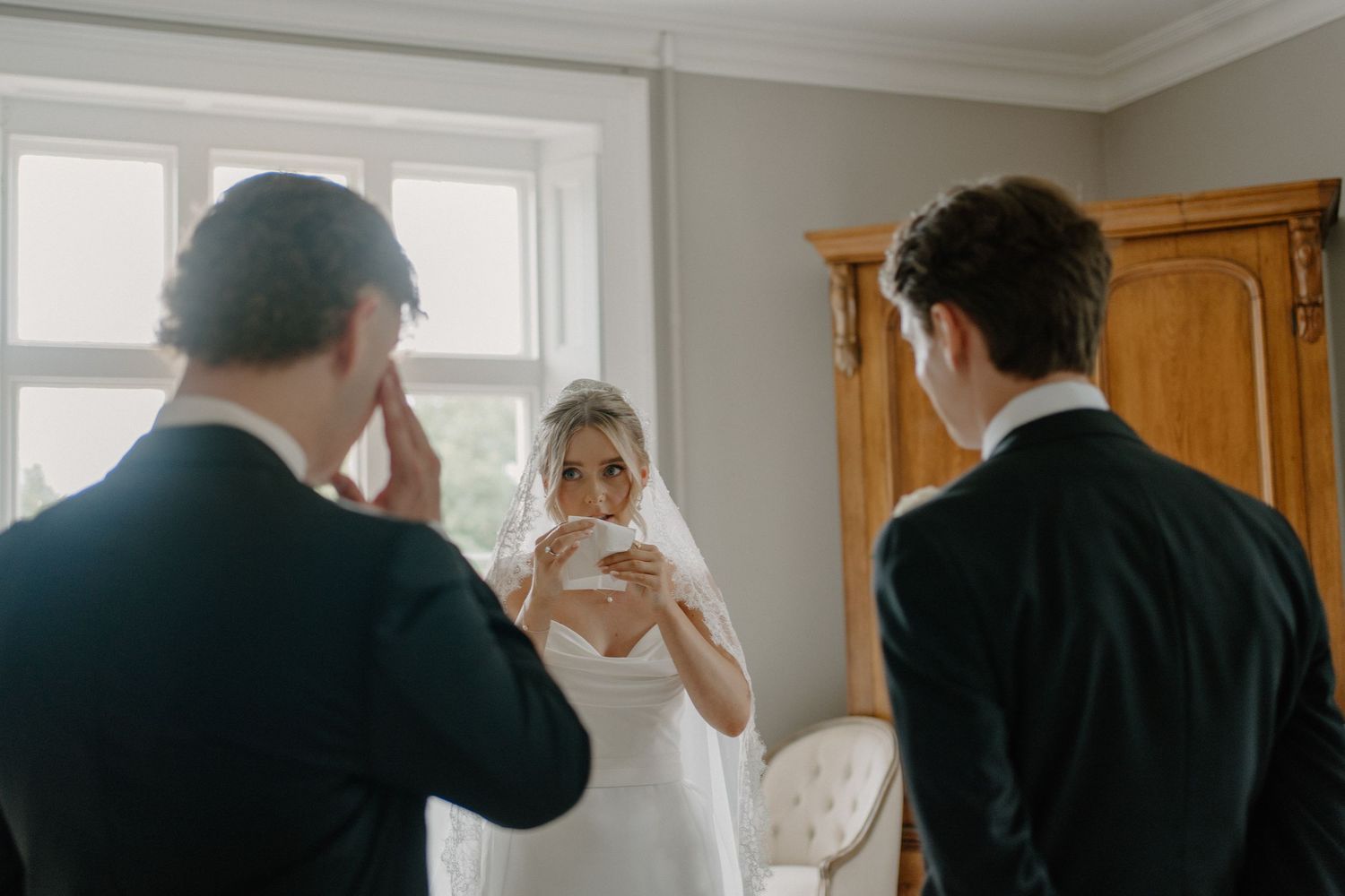 Bride in strapless gown sipping tea while two groomsmen in black suits stand nearby in a bridal suite.