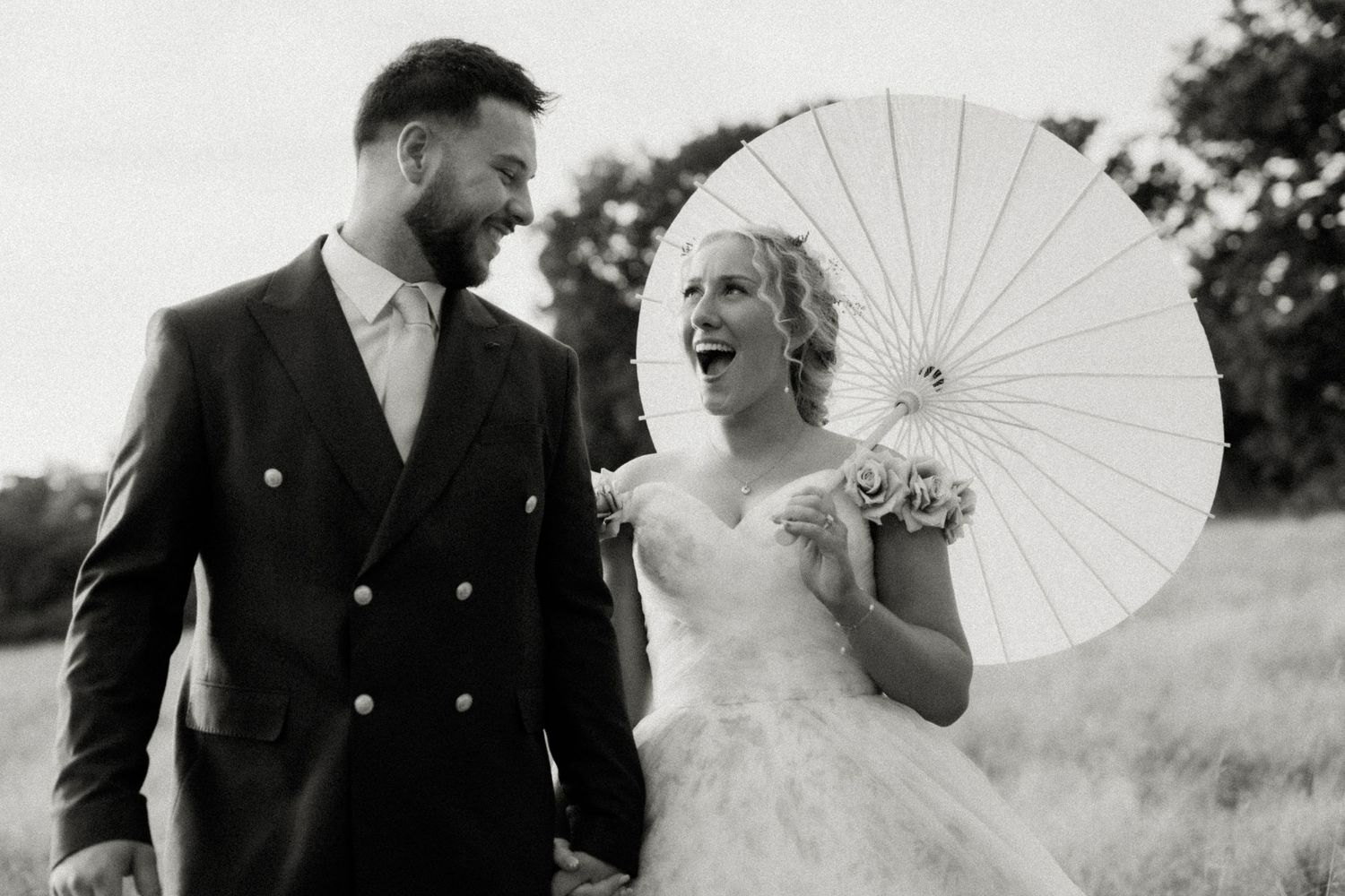 Black and white photo of laughing bride and smiling groom walking together with white parasol outdoors.