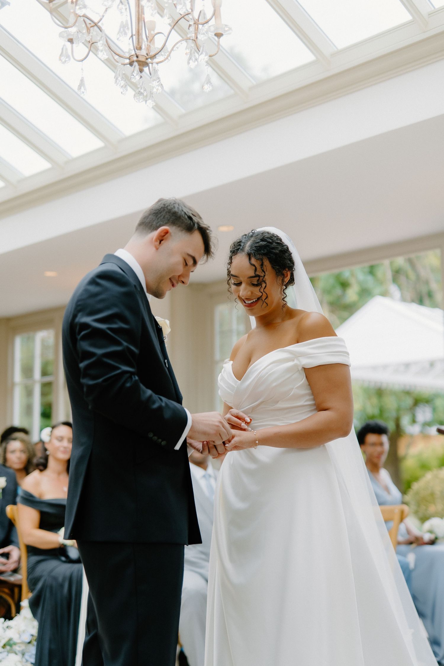 Bride and groom exchange rings during wedding ceremony in elegant glass-roofed venue with chandelier.