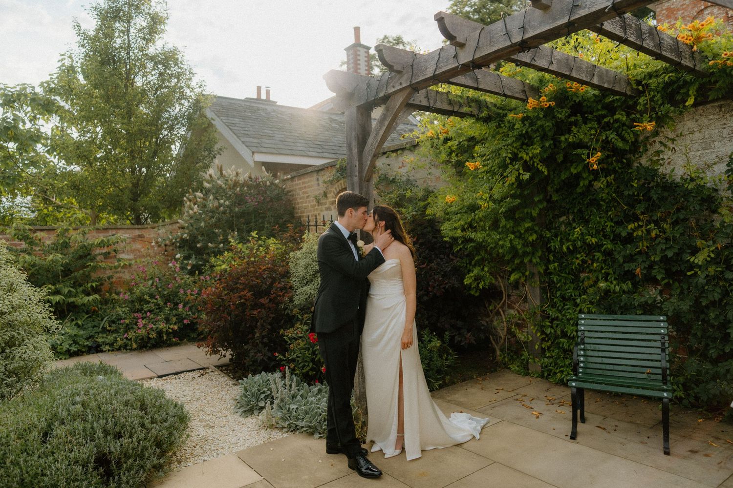 Bride and groom sharing a kiss under a vine-covered pergola in a beautiful garden.